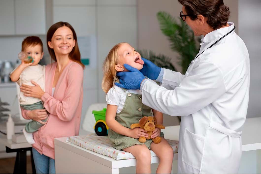 Doctor examining a young girl's throat while a mother and baby watch in a doctor's office.