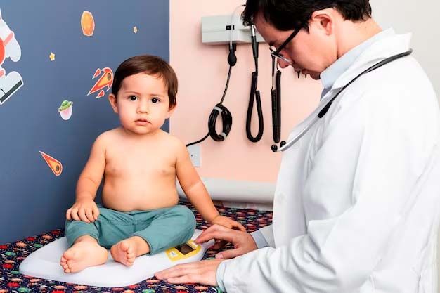 A doctor weighs a shirtless toddler on a scale in an exam room.