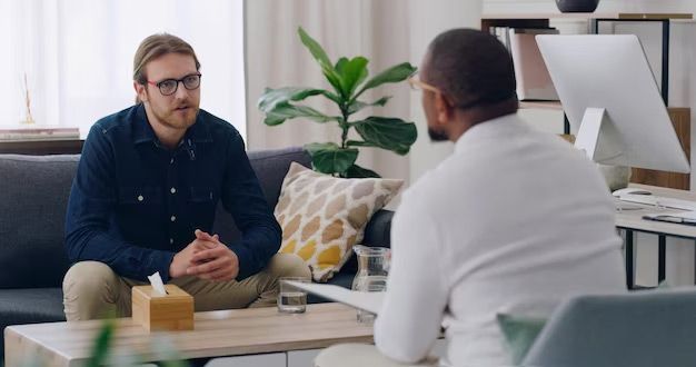 Man with glasses talking to therapist in a light-filled room. Therapist takes notes; tissue box on table.