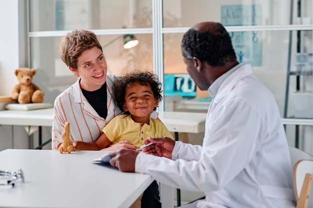 A doctor consults with a mother and child in a bright office. The child smiles, and the mother looks on.