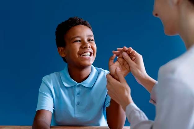 Smiling boy in blue shirt looks at woman gesturing with hands in front of a blue background.