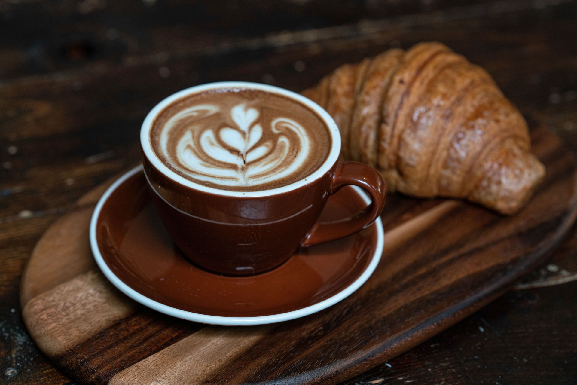 Una tazza marrone di latte macchiato con una decorazione a forma di cuore sul piattino, accanto a un croissant su un vassoio di legno.