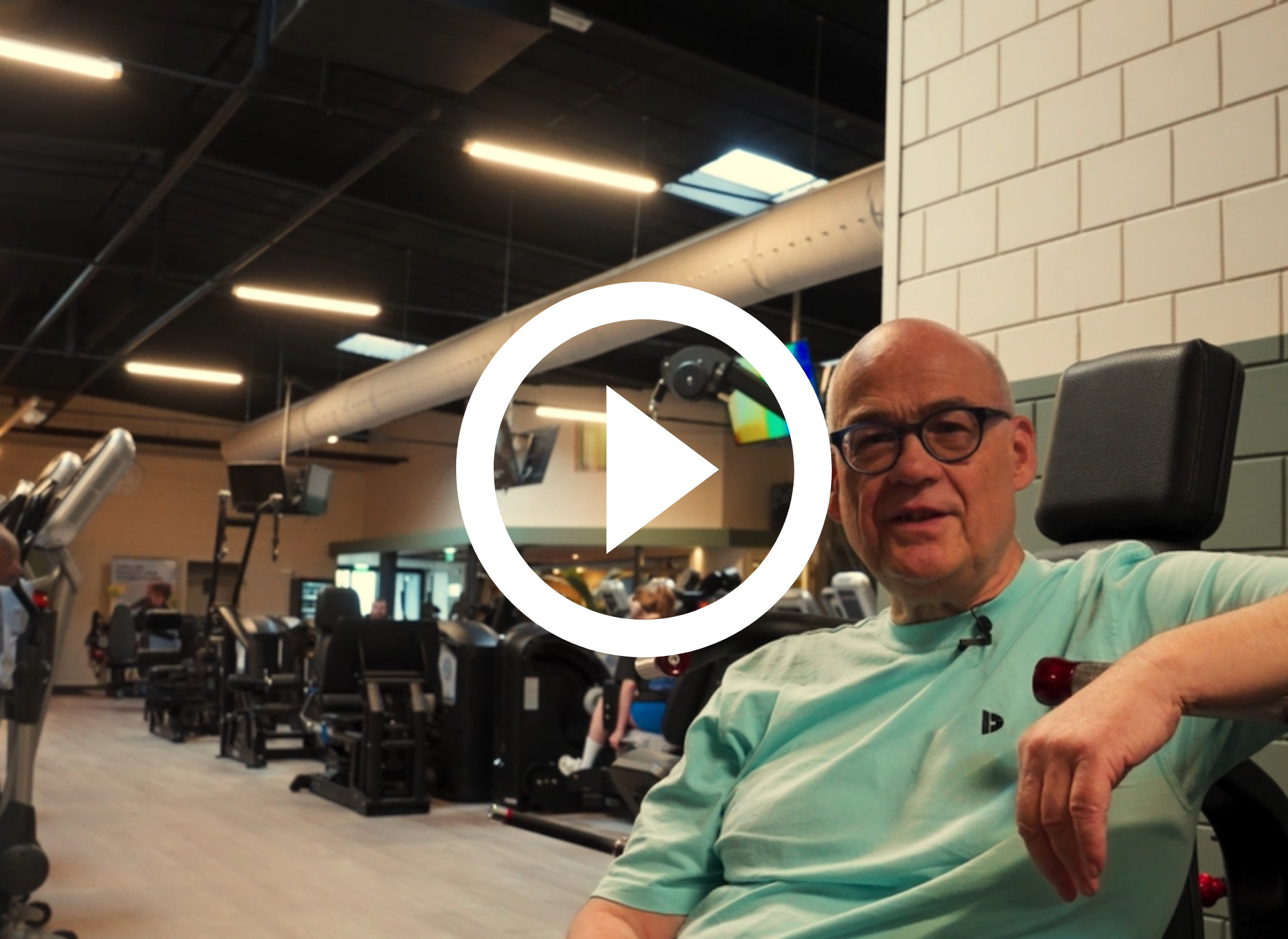 Een man zit in een sportschool, draagt een bril en een mintgroen shirt, met fitnessapparaten op de achtergrond.