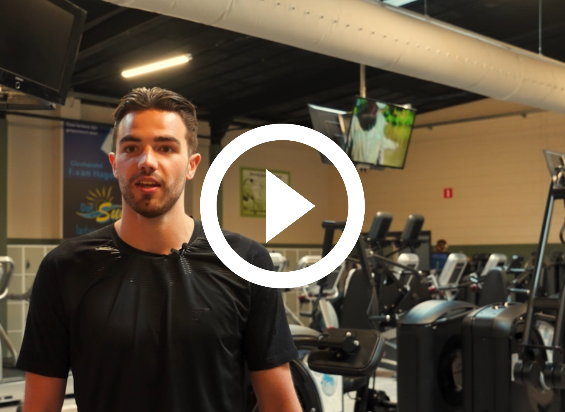 Een man staat in een sportschool naast fitnessapparaten, met een afspeelknop als overlay op de afbeelding.