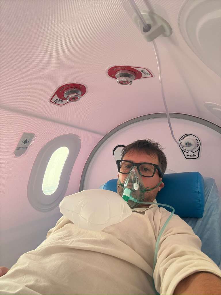 Un hombre yace en un entorno médico, con una mascarilla de oxígeno, mirando a la cámara.