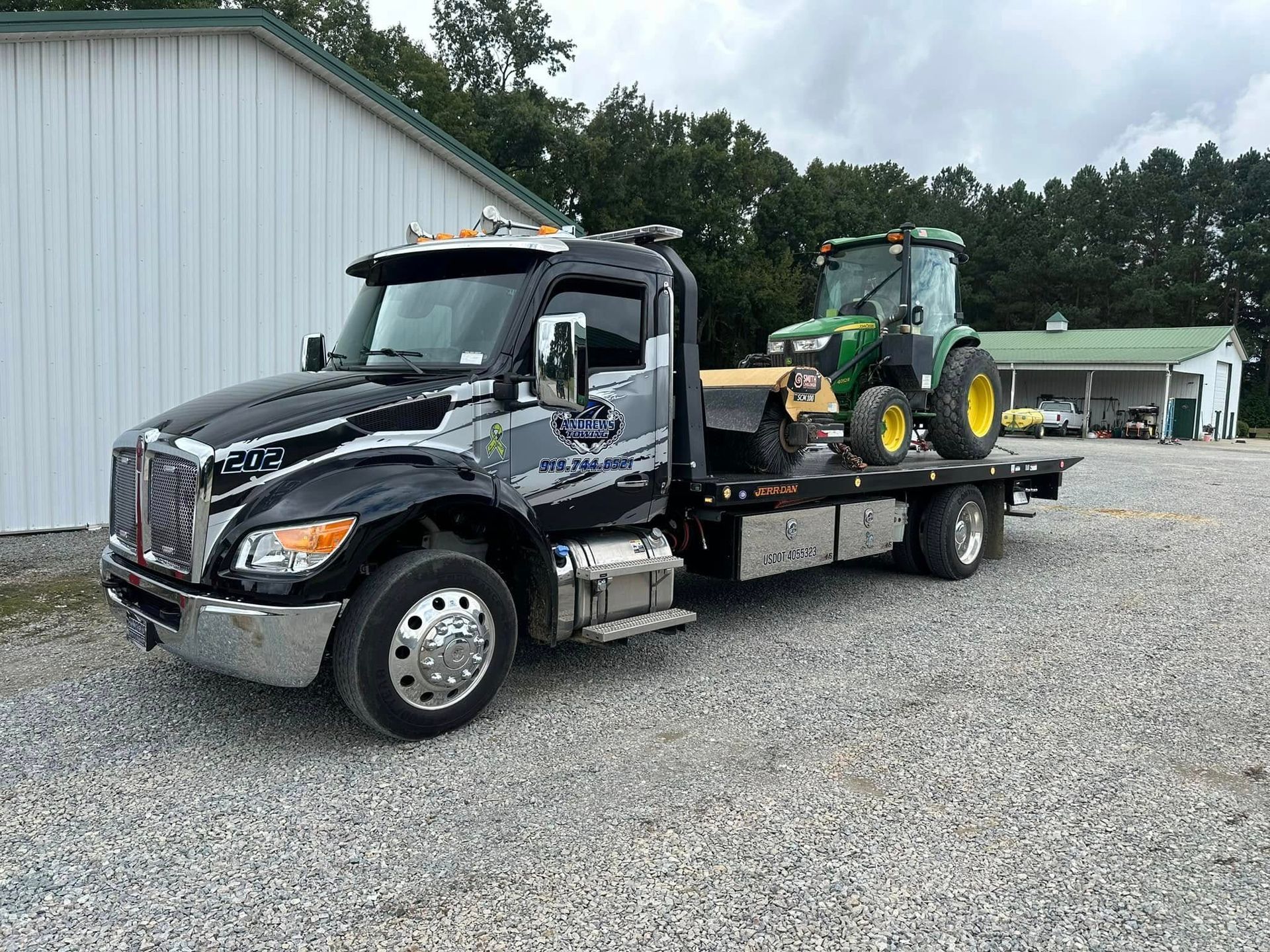 A tow truck with a tractor on the back is parked in a gravel lot.