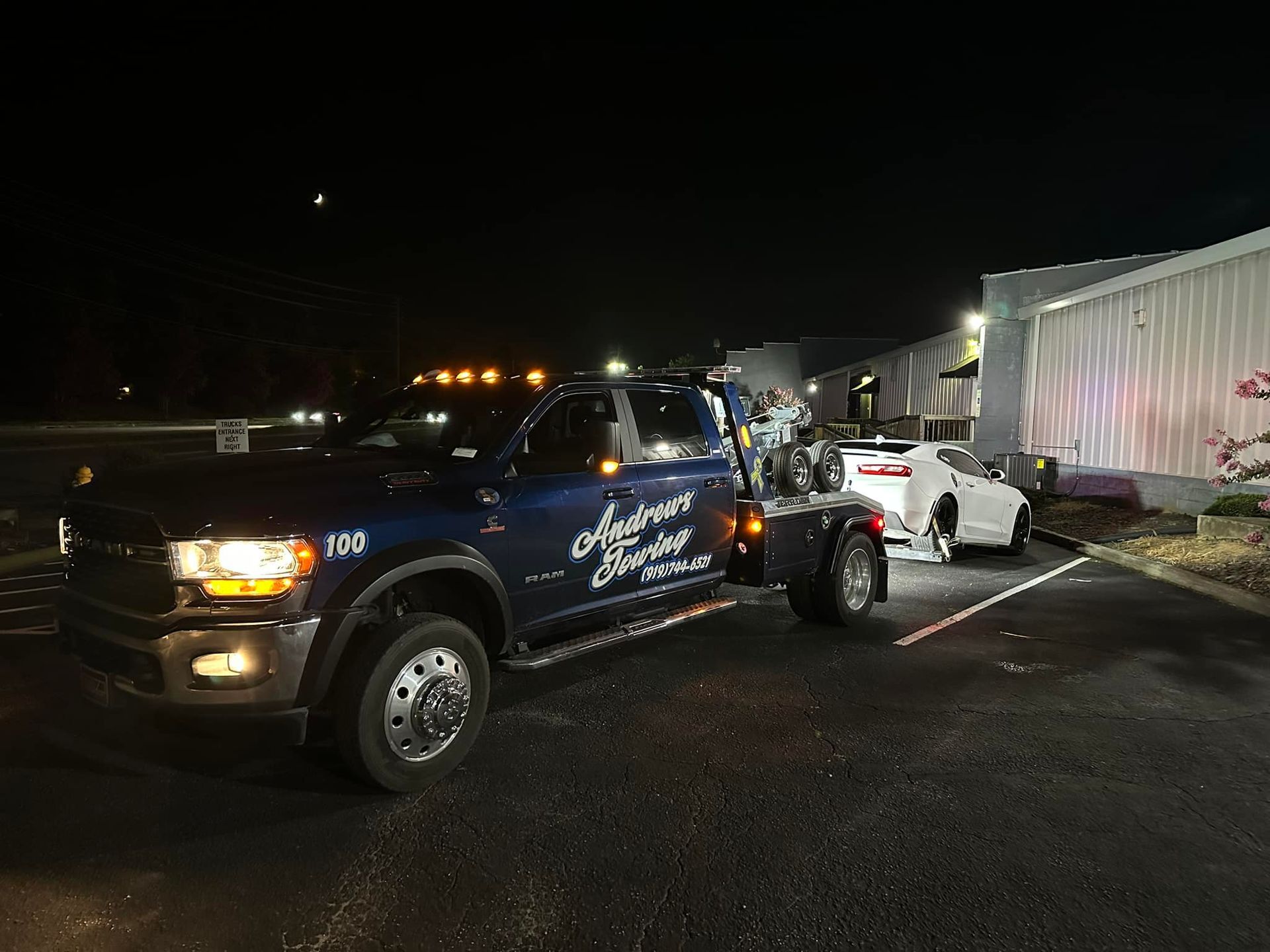 A blue tow truck is towing a white car in a parking lot at night.