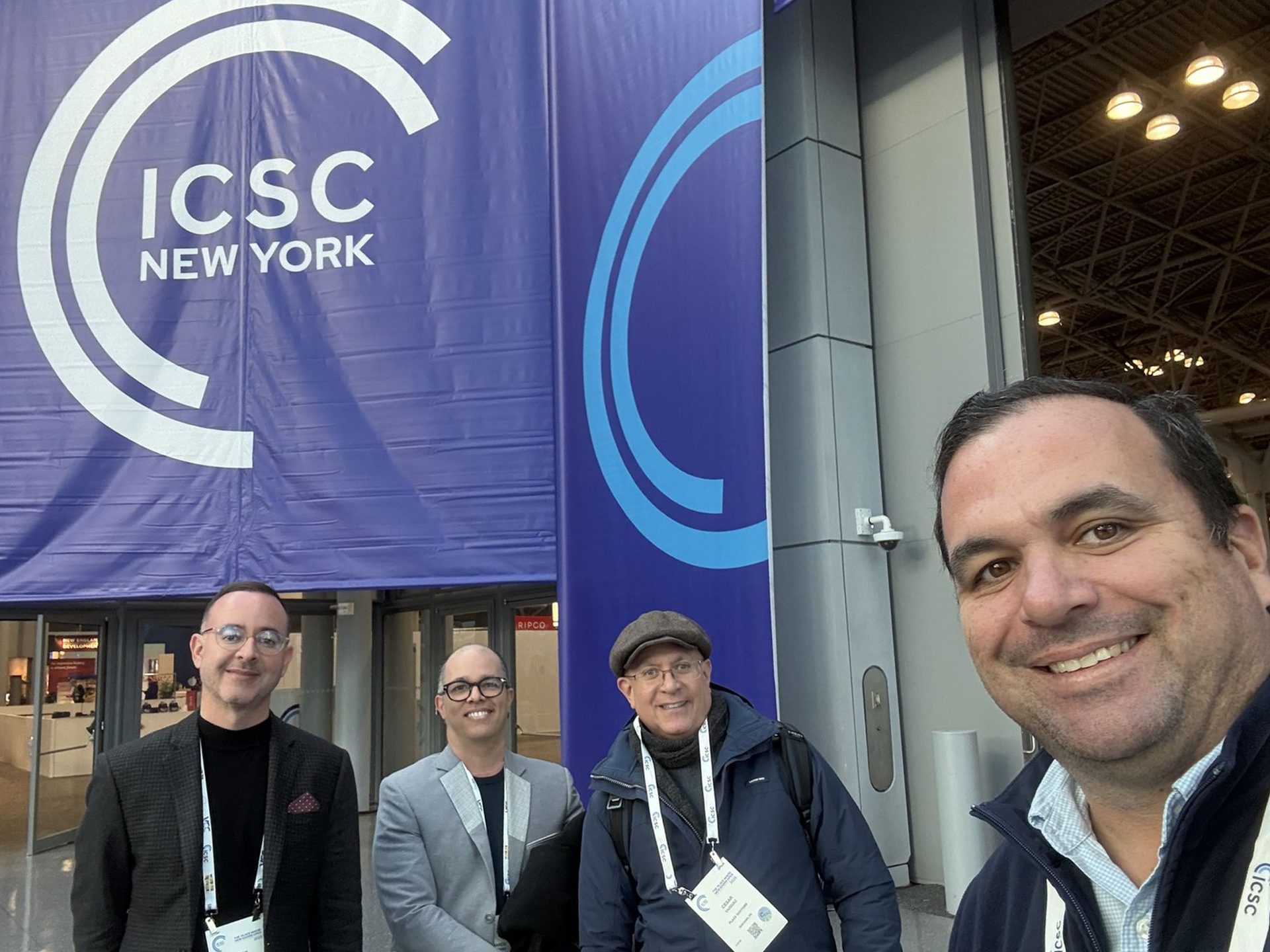 Four people at ICSC New York event. Men in front of entrance with large banner, smiling and looking at camera.