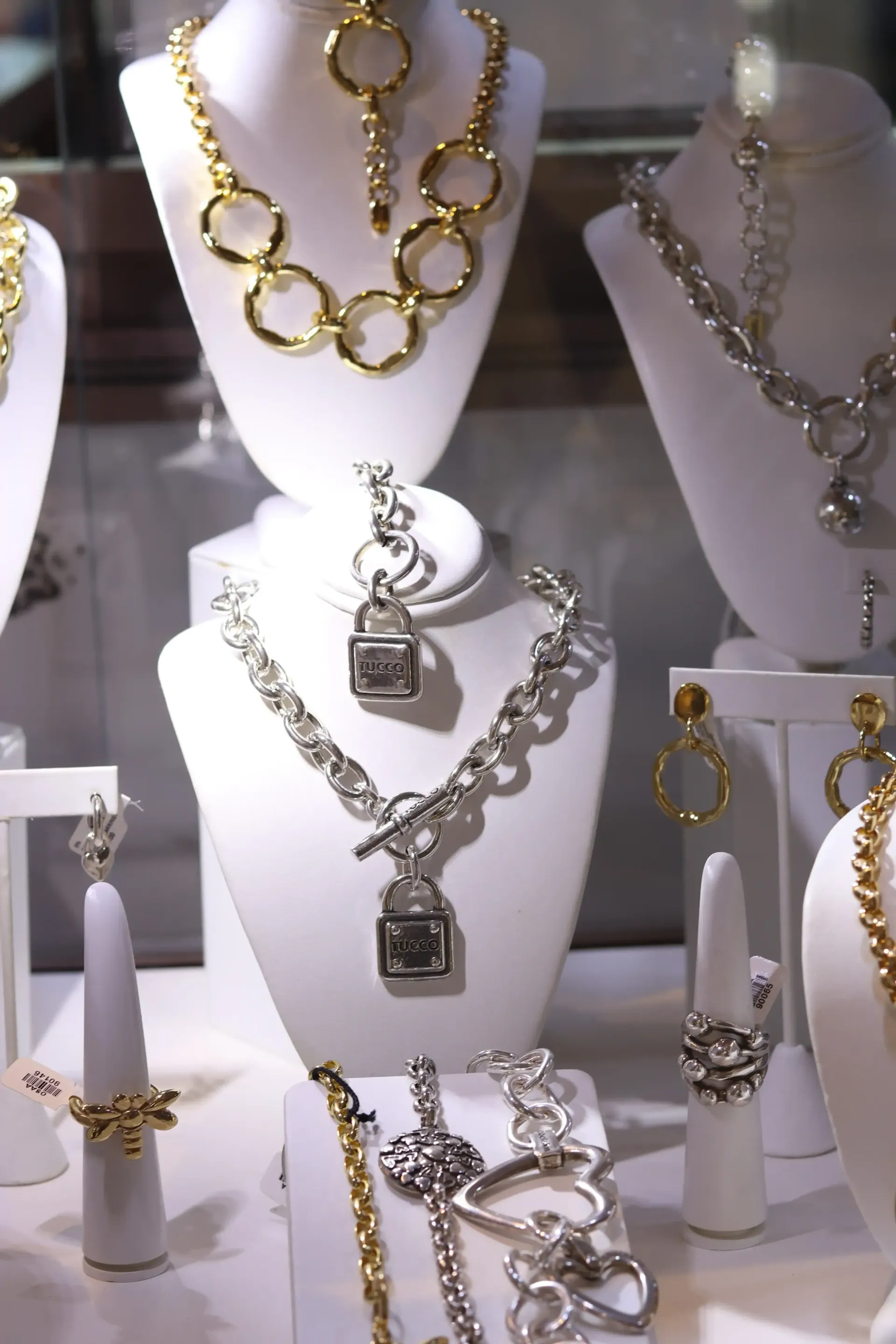 Jewelry display with gold and silver necklaces, bracelets, and rings on white mannequins, inside a shop window.