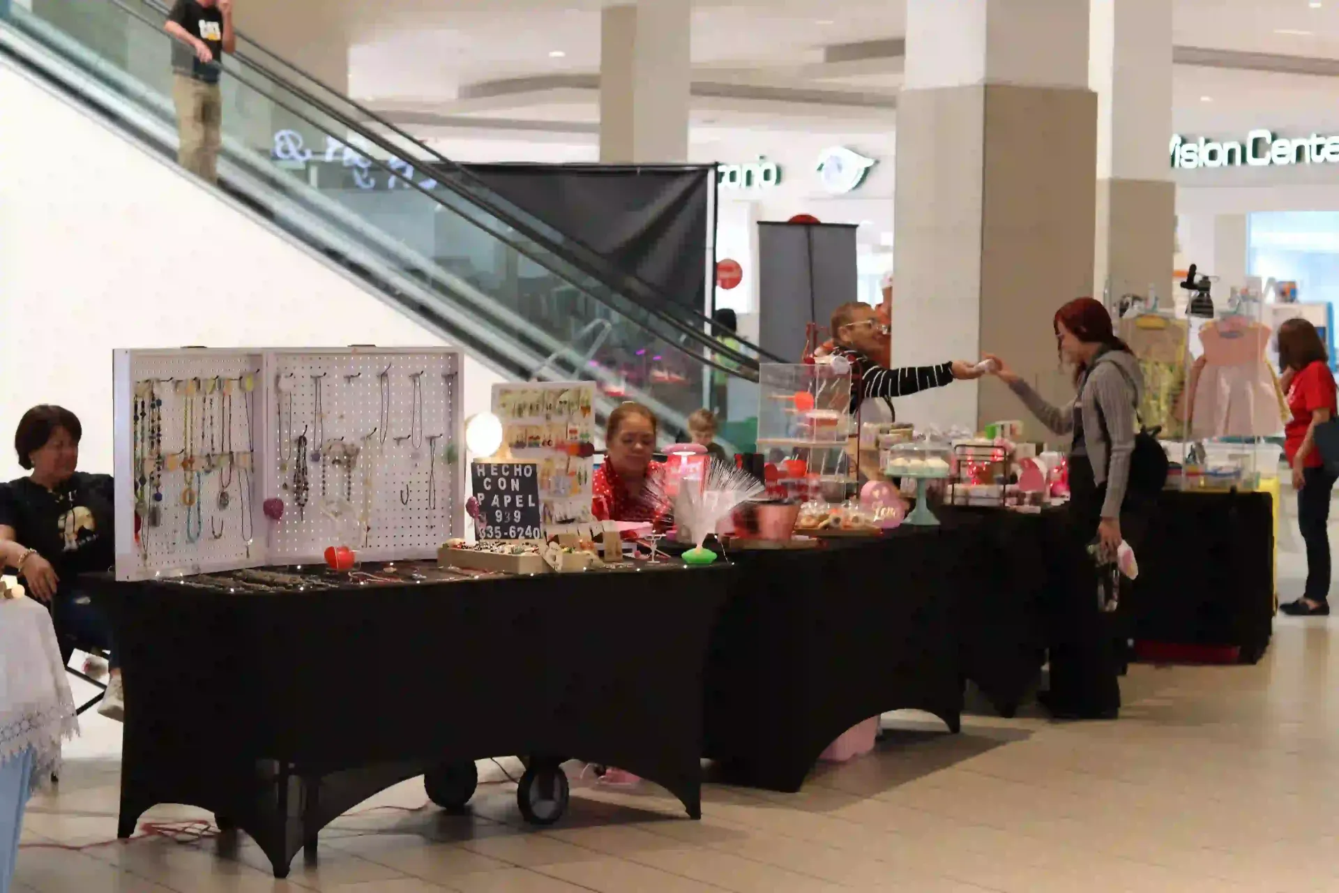 People browsing craft tables at an indoor event, with jewelry, baked goods, and dresses displayed.