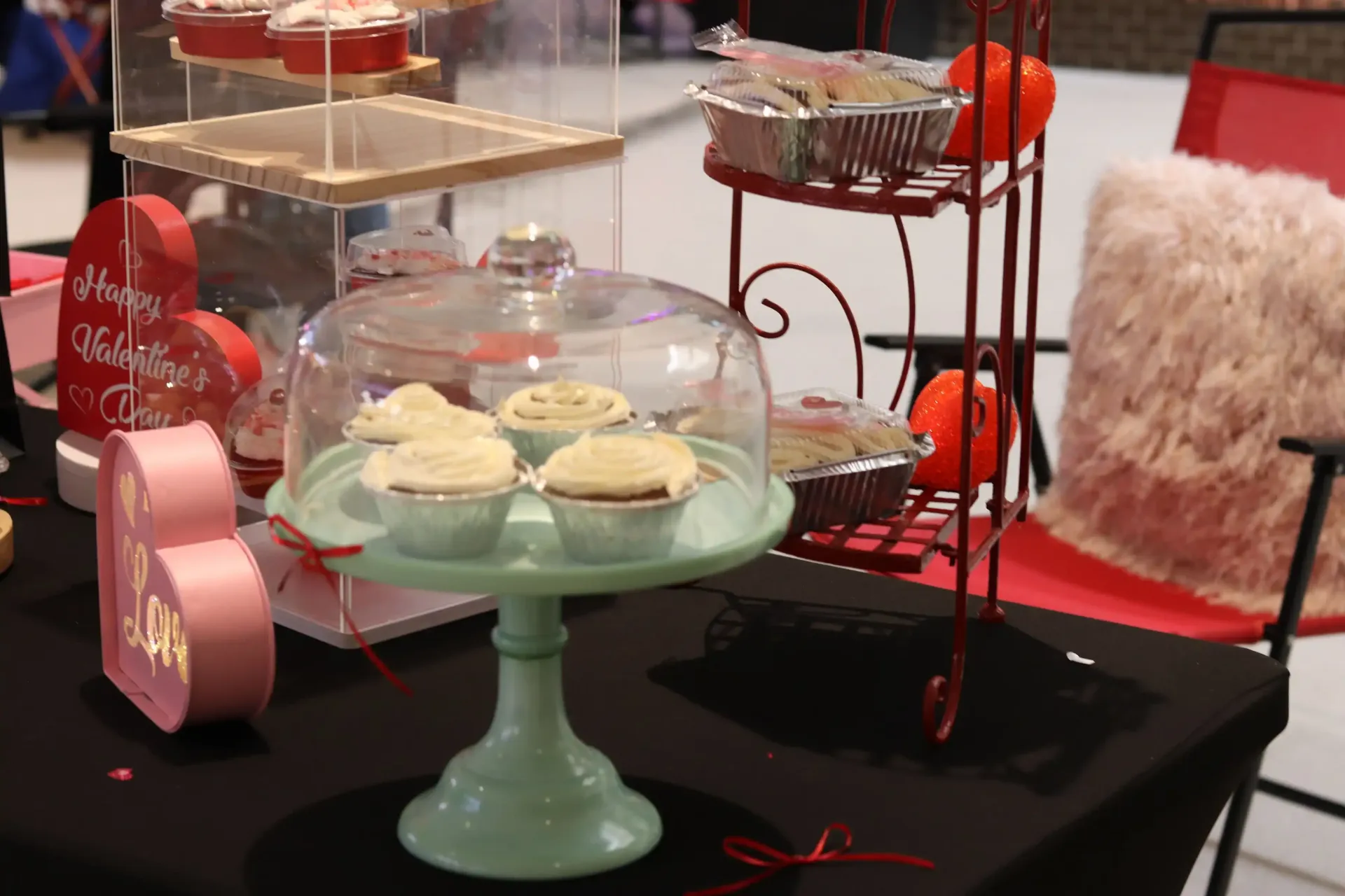 A display of cupcakes under a glass dome, with other baked goods, and Valentine's decorations on a table.
