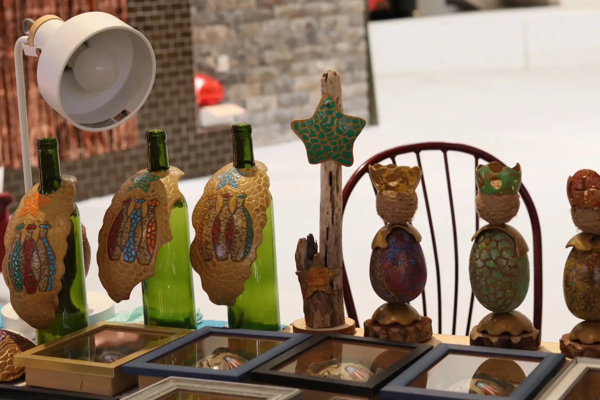 Display of decorated bottles and egg-shaped art pieces. A white lamp and brick background are also visible.