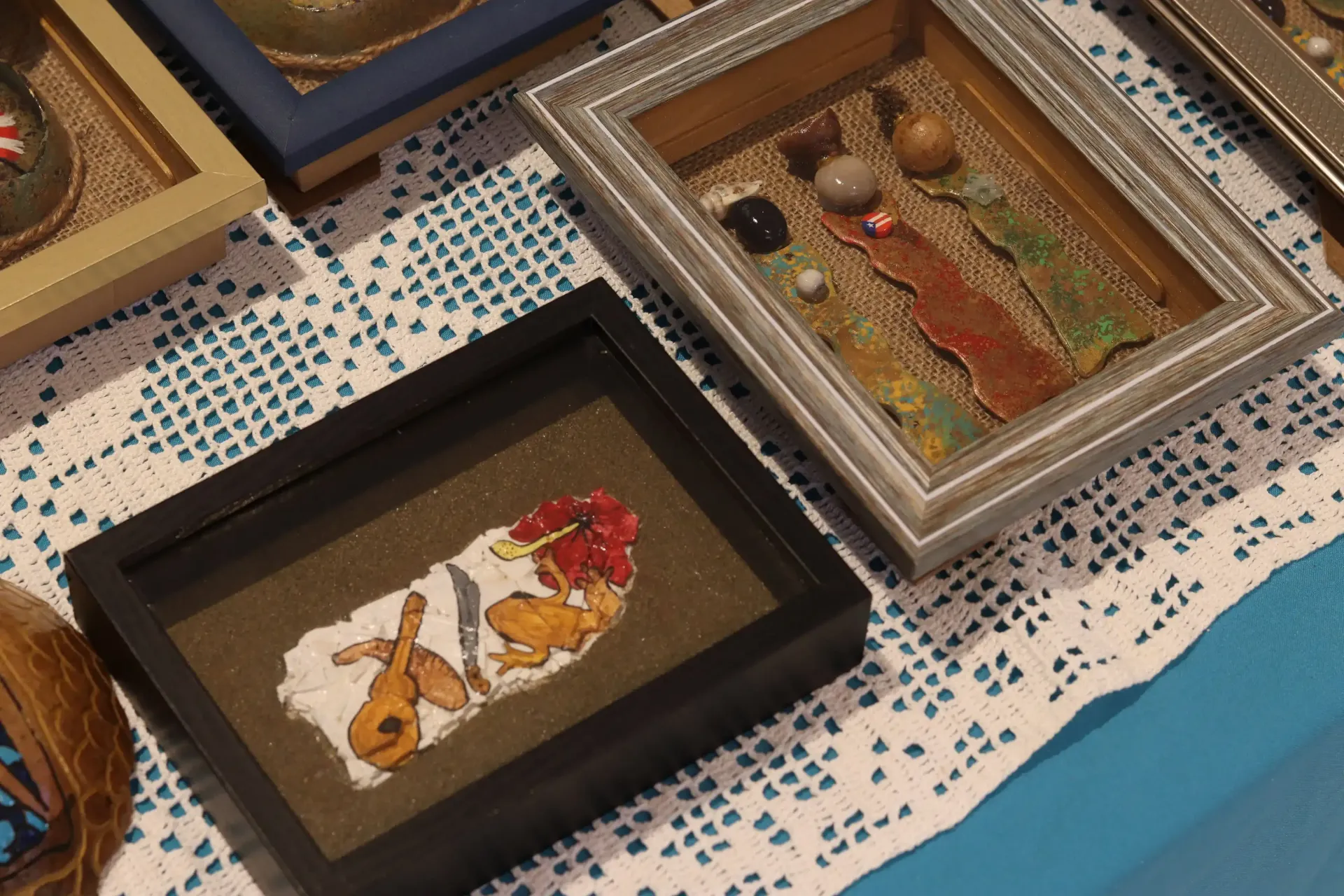 Framed craft art with various objects, including rocks, colored shapes, and a guitar. Set on a white lace cloth.