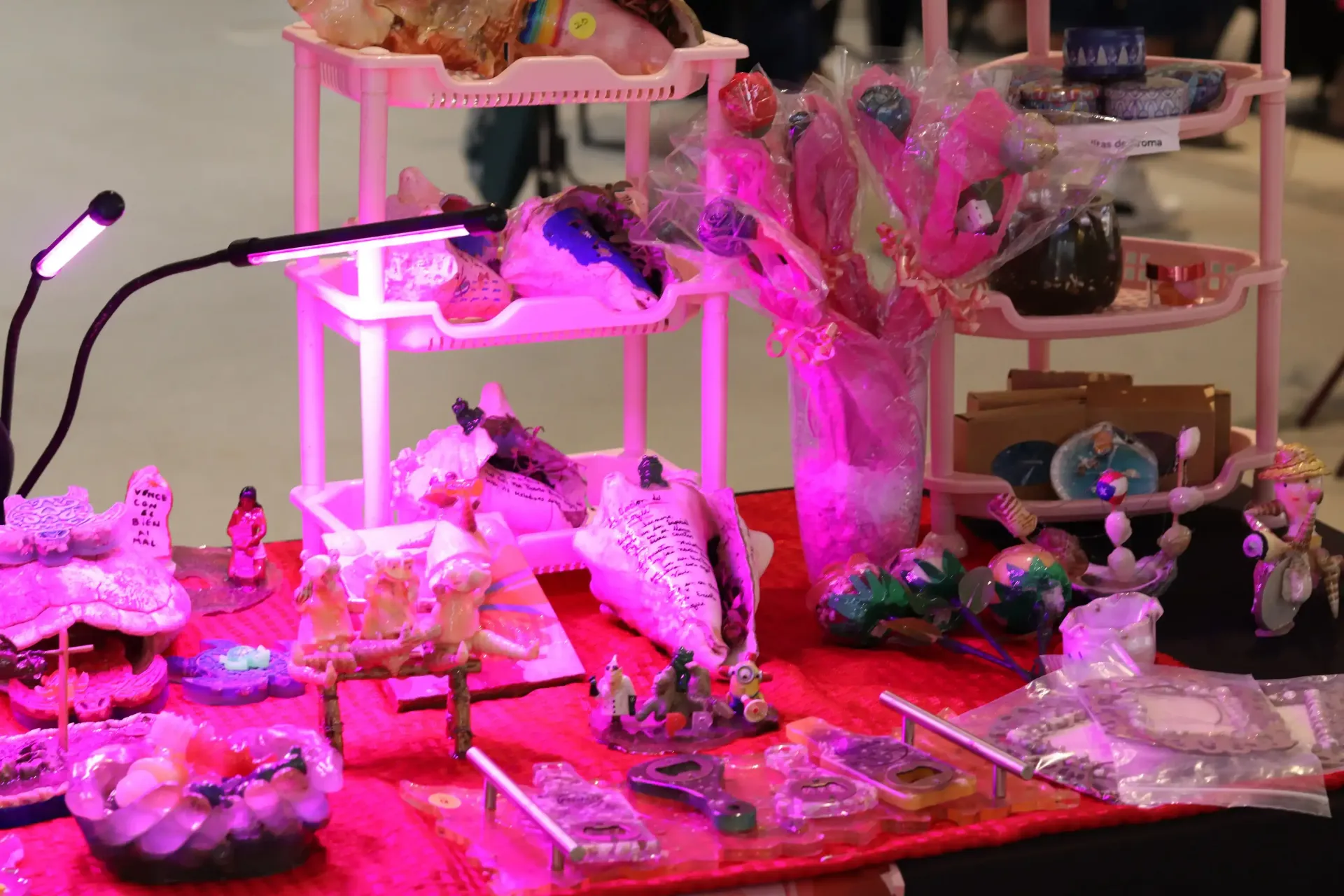 A display of colorful crafts, jewelry, and gifts lit by a pink light at a craft fair. Items are on multiple tiers of shelves.