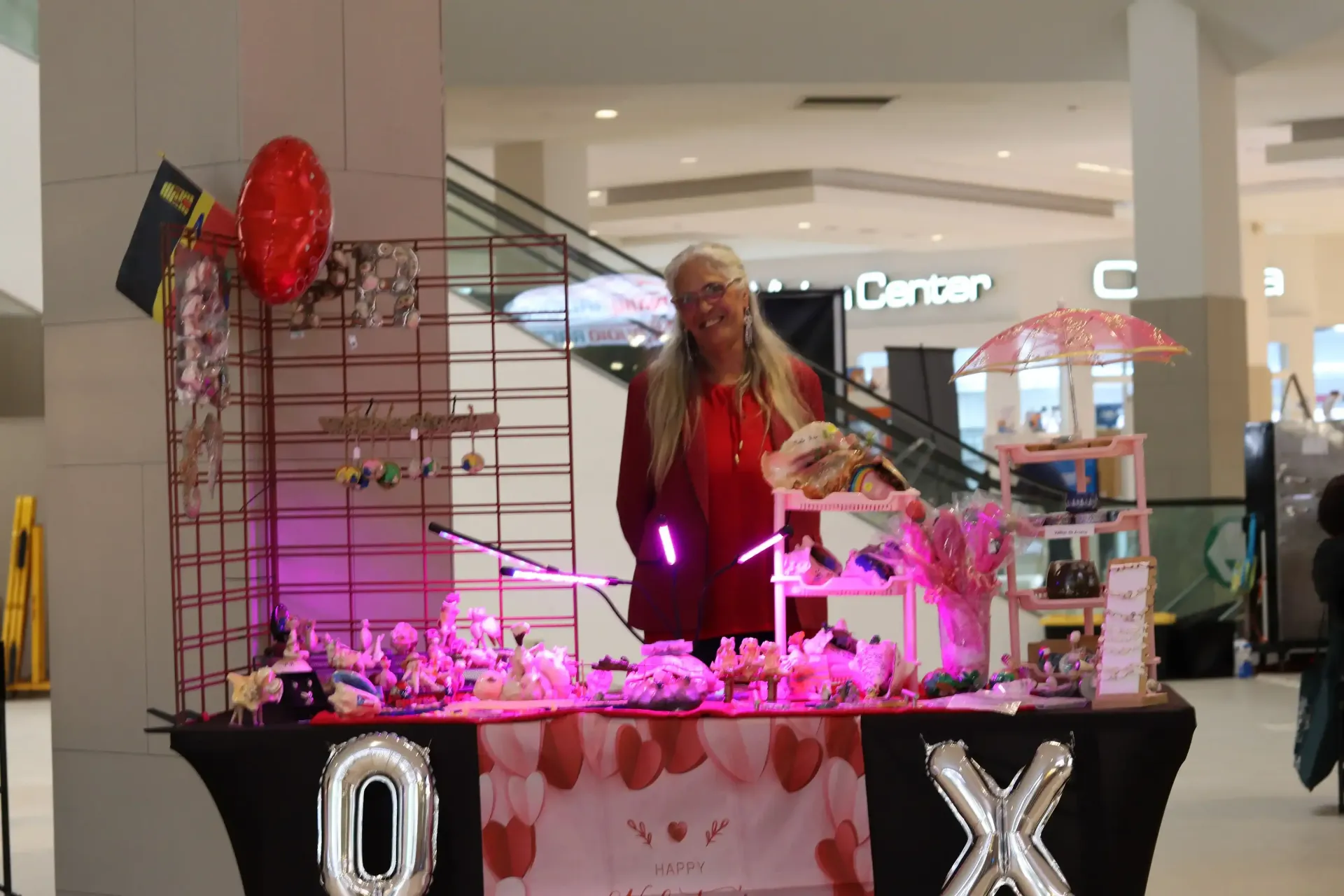 Woman selling Valentine's Day themed jewelry and decorations at a mall kiosk, lit with pink lights.