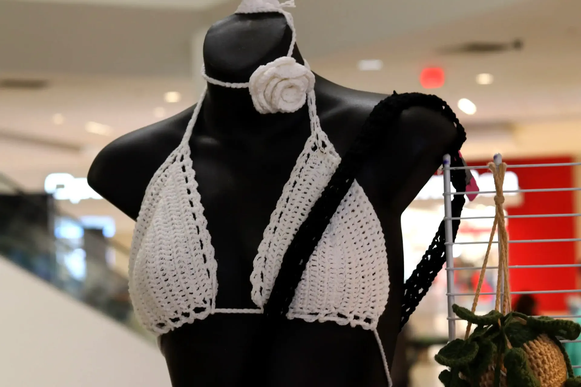 Black mannequin wearing a white crochet bikini top with a matching flower choker. Shopping mall in the background.