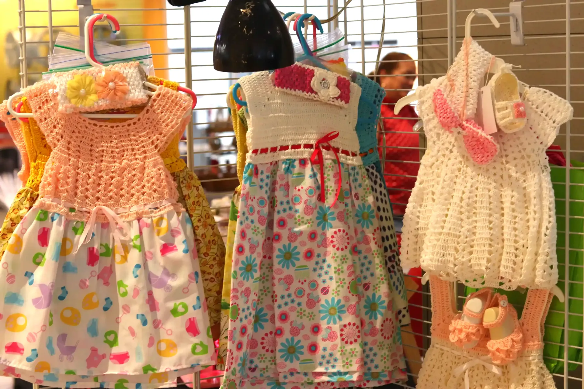 Crocheted baby dresses in peach, white, and cream with matching headbands and shoes, displayed on hangers.