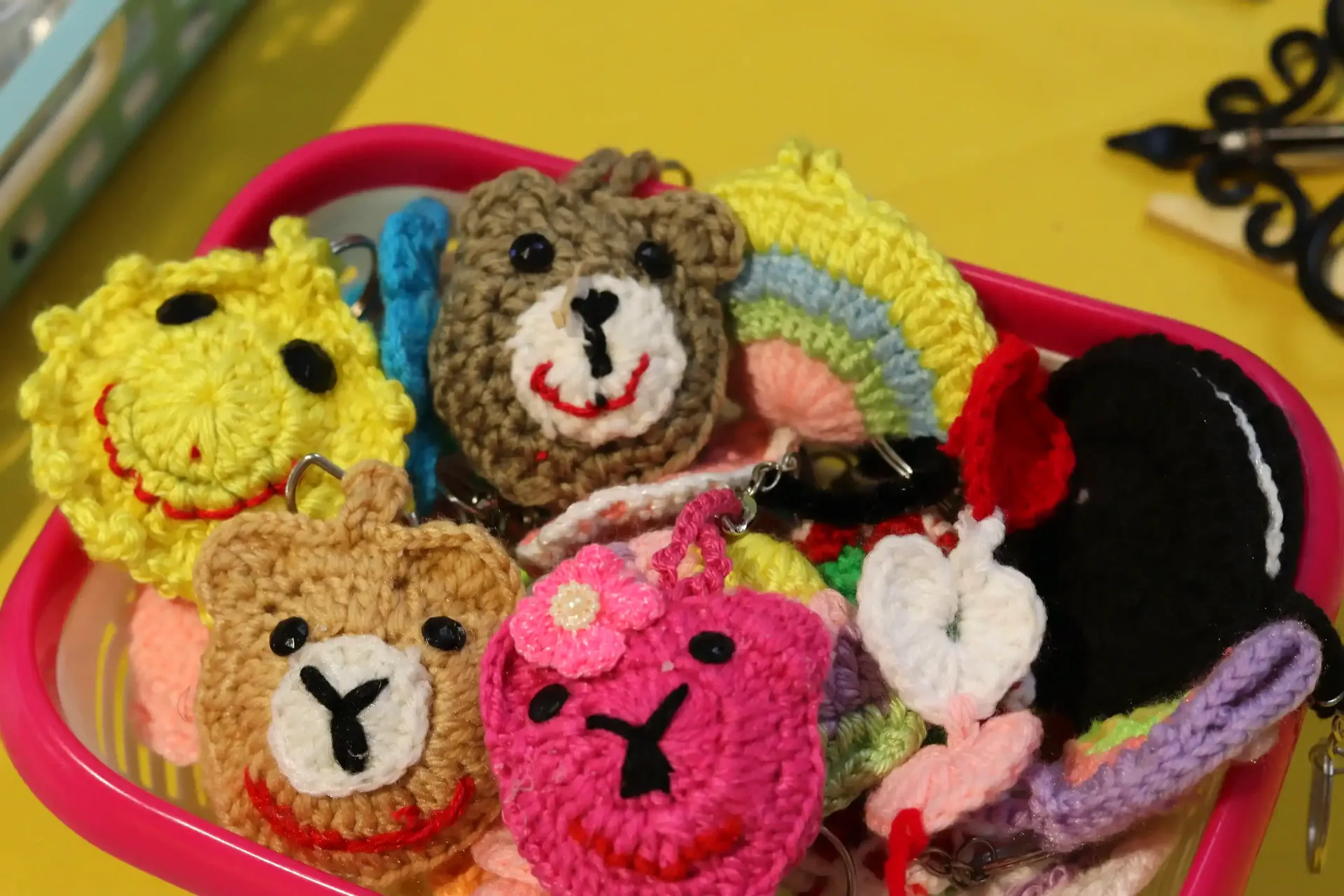 A pink basket overflowing with colorful, crocheted animal keychains, including bears and a sun, on a yellow surface.