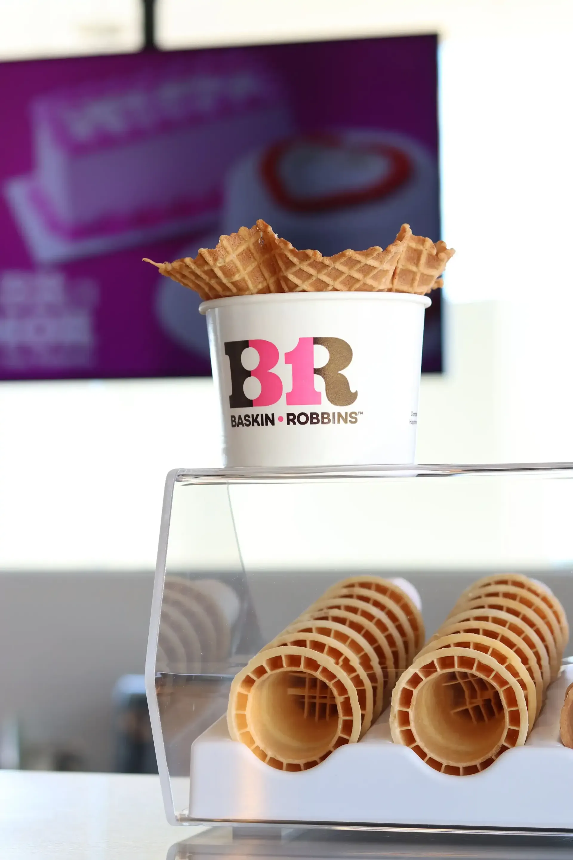 A Baskin-Robbins cup with waffle cone tops and display case filled with cones in a brightly lit setting.