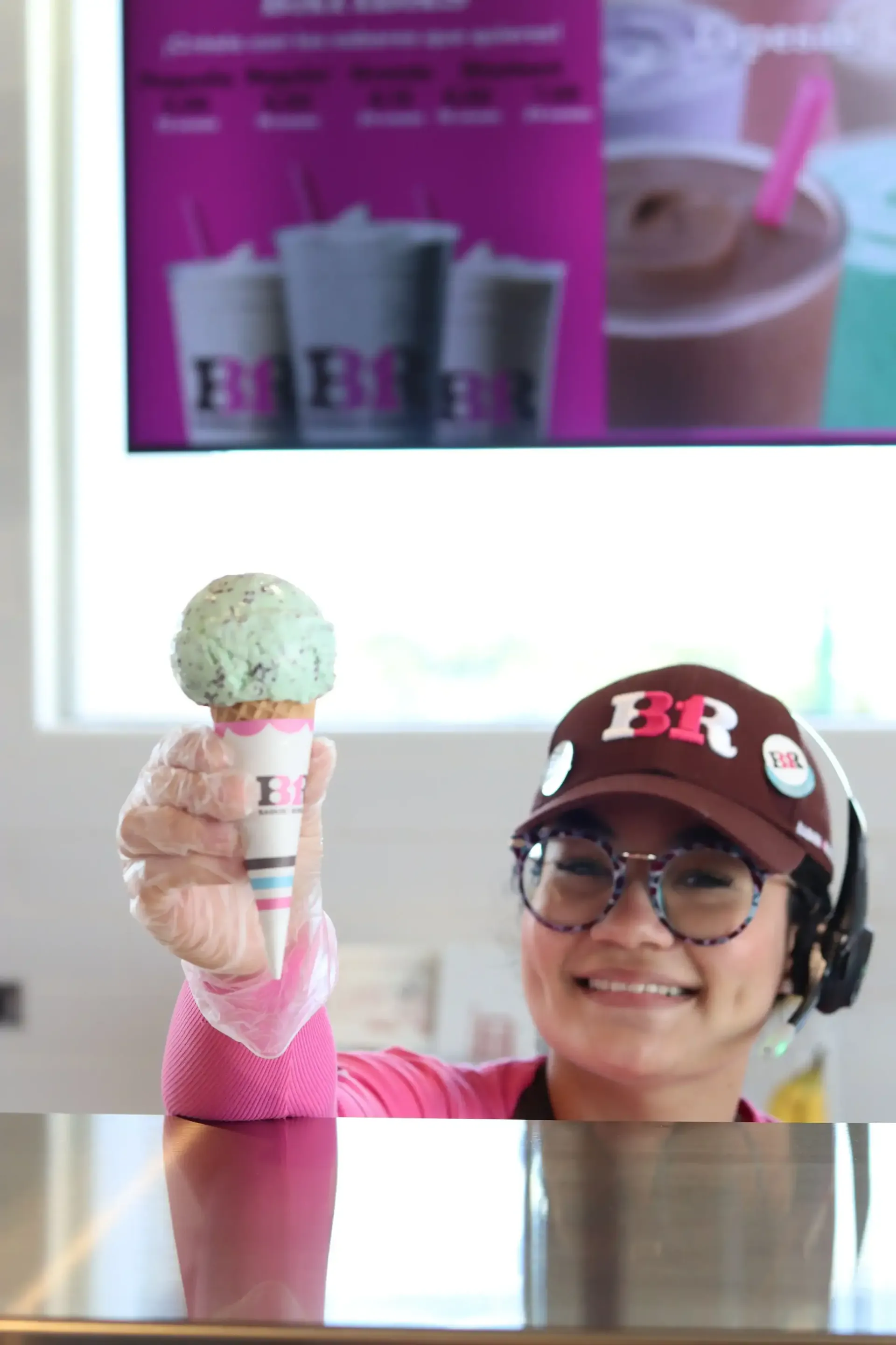 A Baskin-Robbins employee smiling and holding up a mint chocolate chip ice cream cone. She wears a cap and glasses.