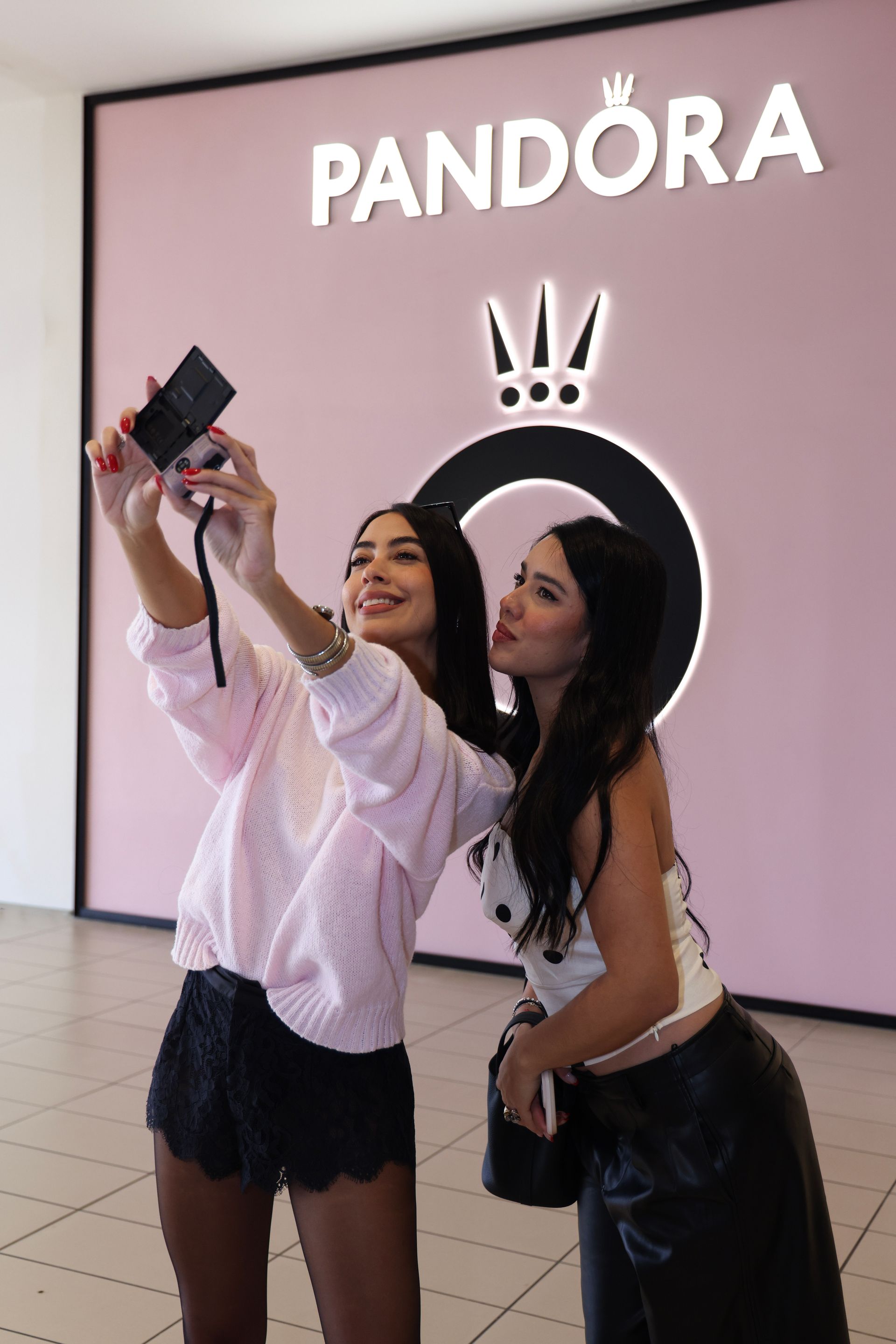 Two women taking a selfie in front of a PANDORA sign and logo display.