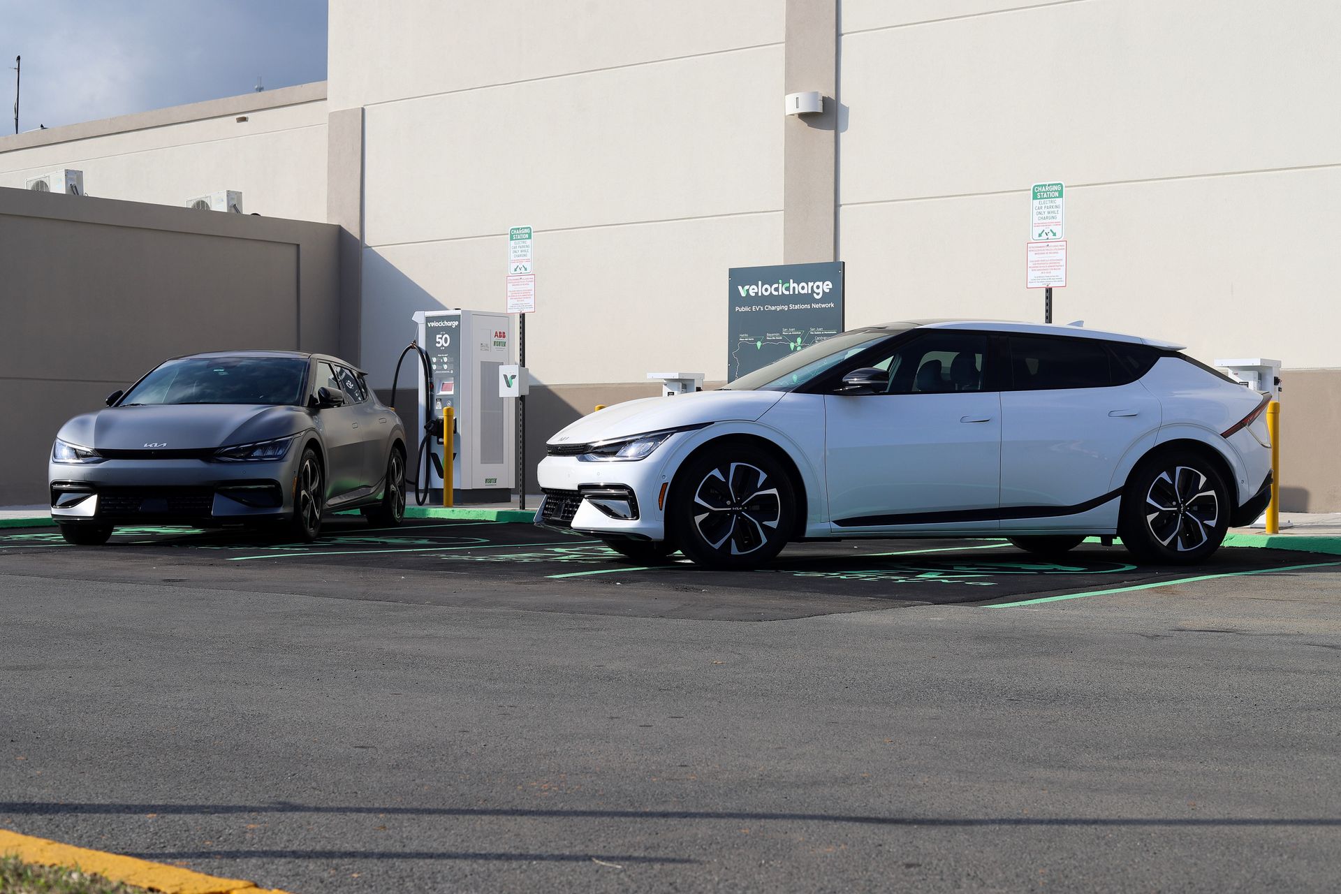 Two Kia EVs charging at a station, one gray, one white. Outdoor setting.