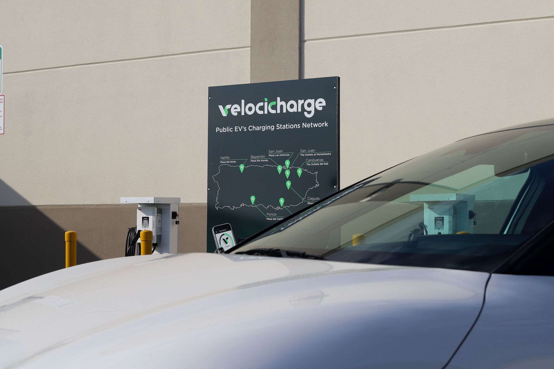 White car at a Velocharge EV charging station; sign and charging ports are visible.
