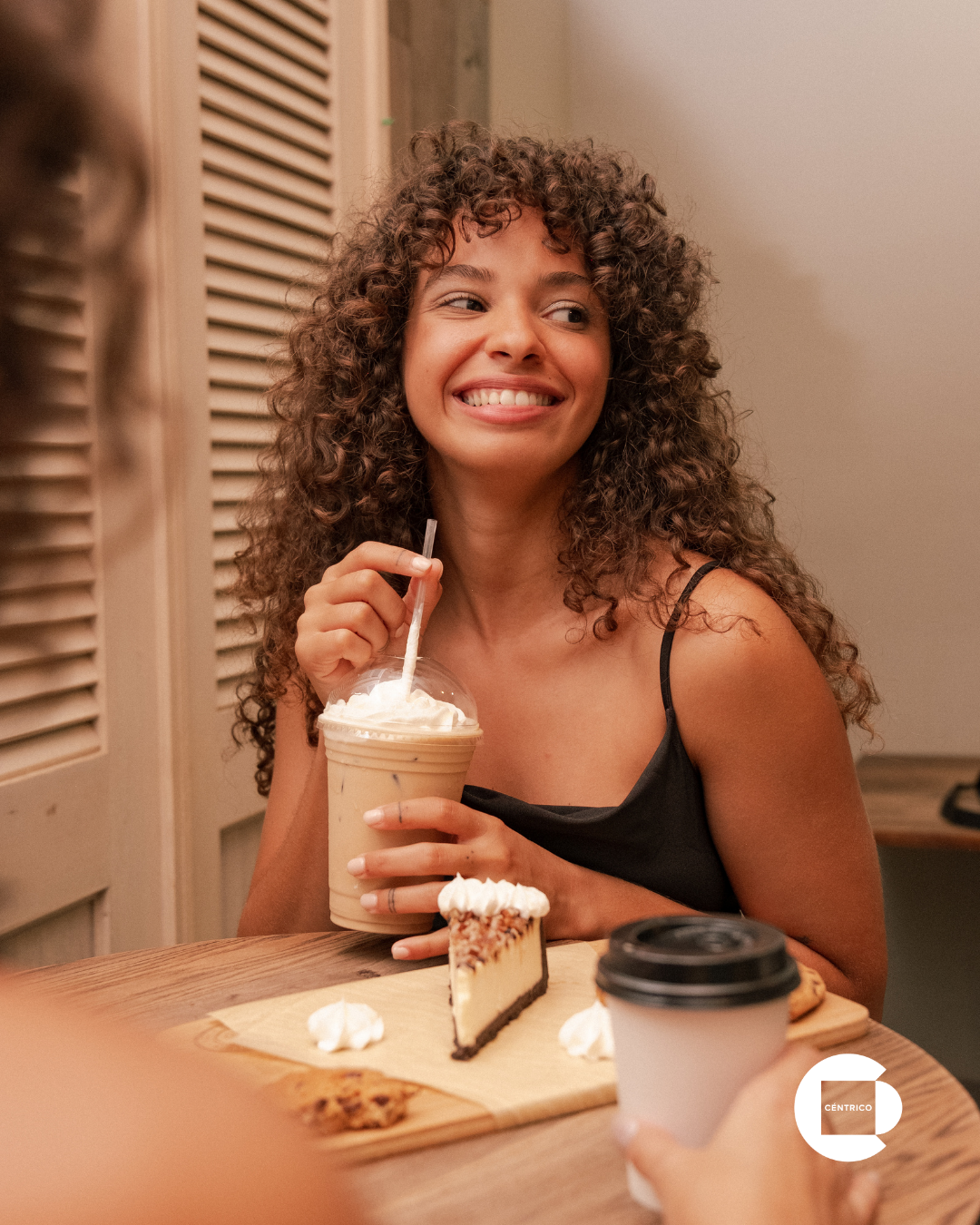 A curly-haired person smiling at a companion while holding an iced coffee at a cafe table with a slice of cheesecake.