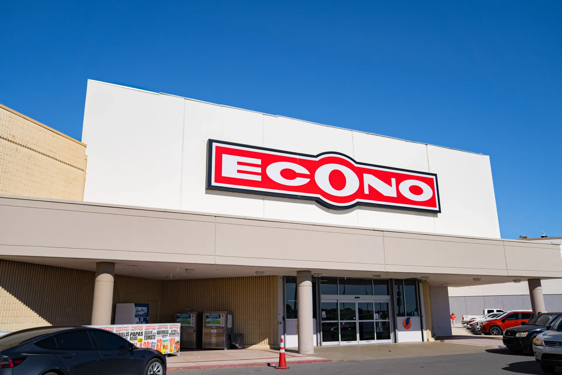 Econo grocery store entrance under a bright blue sky, with a red and white logo. Cars are parked in front.