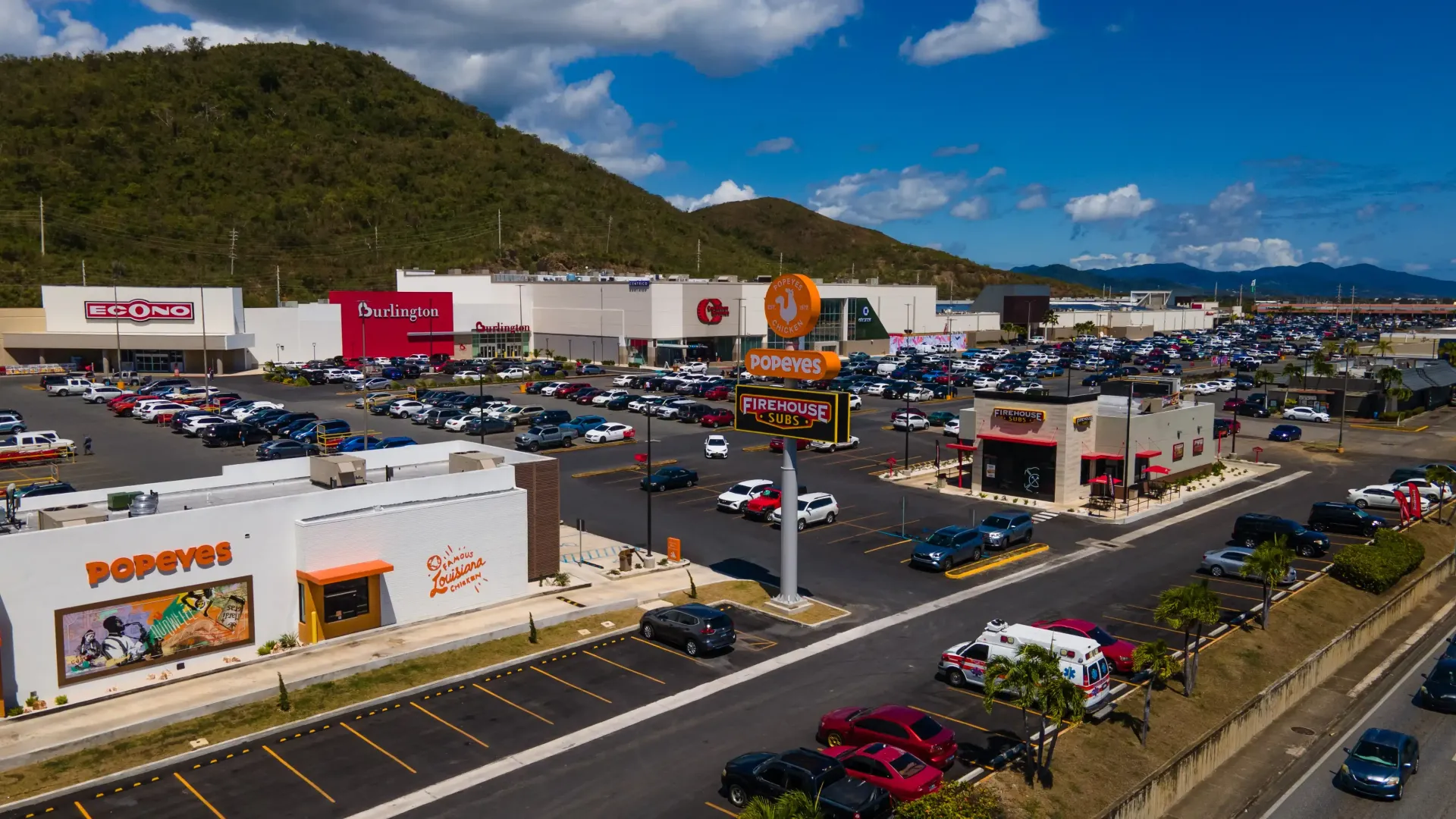 Aerial view of Céntrico Guayama a shopping center with a large parking lot, restaurants (Popeyes), and various stores.