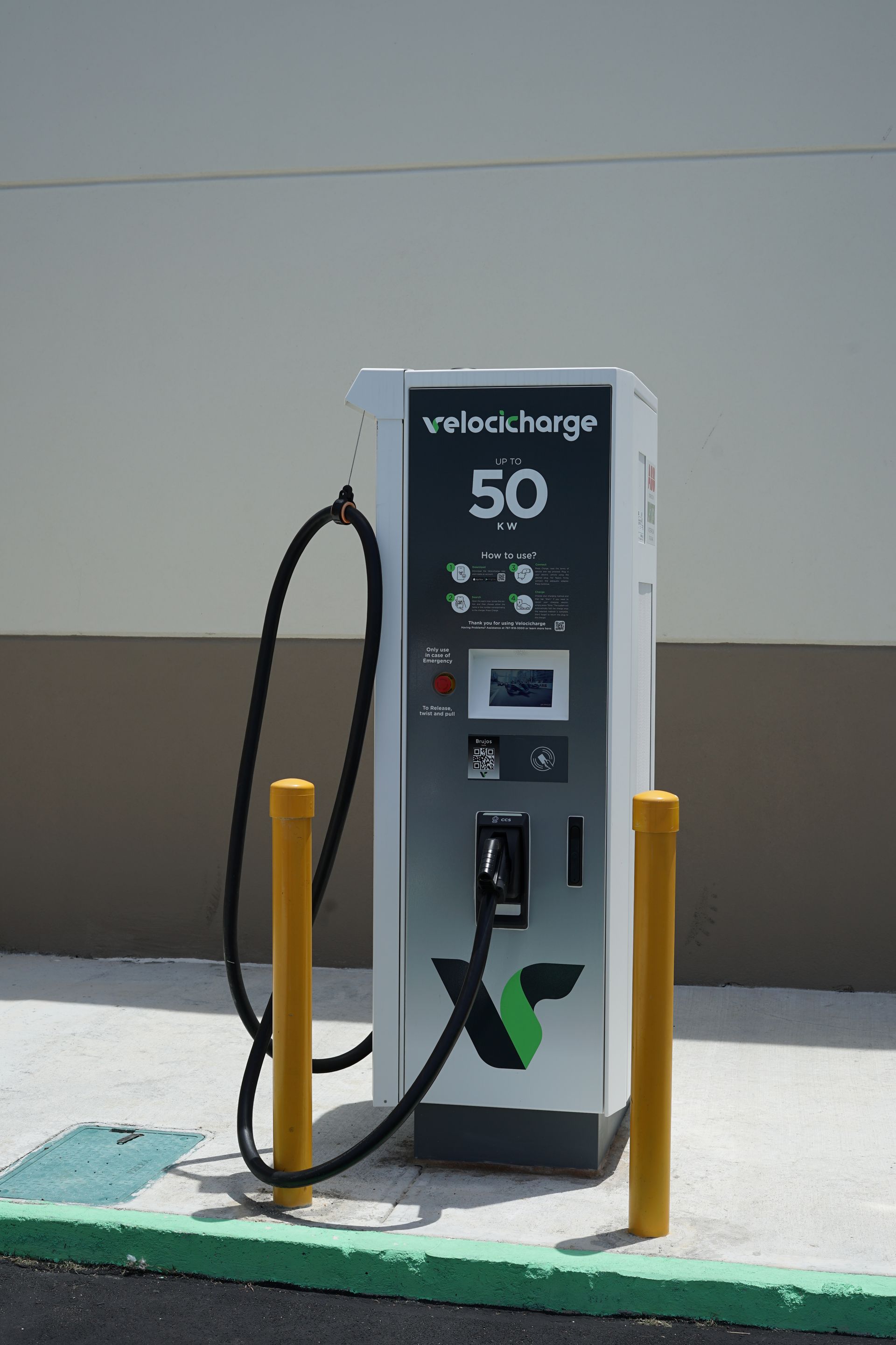 Electric vehicle charging station; white and silver with green logo, black cable and yellow posts.