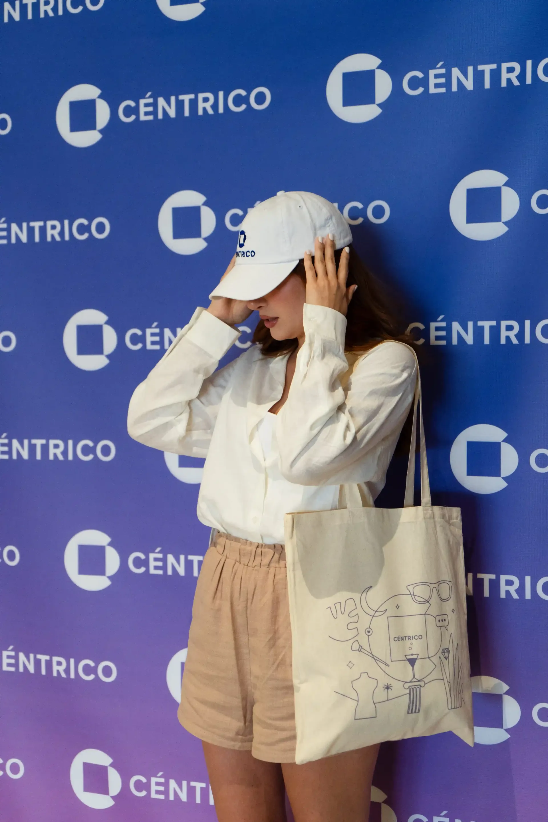 A young woman in a white shirt, tan shorts, and a white hat poses in front of a blue Centrico backdrop, holding a tote bag.