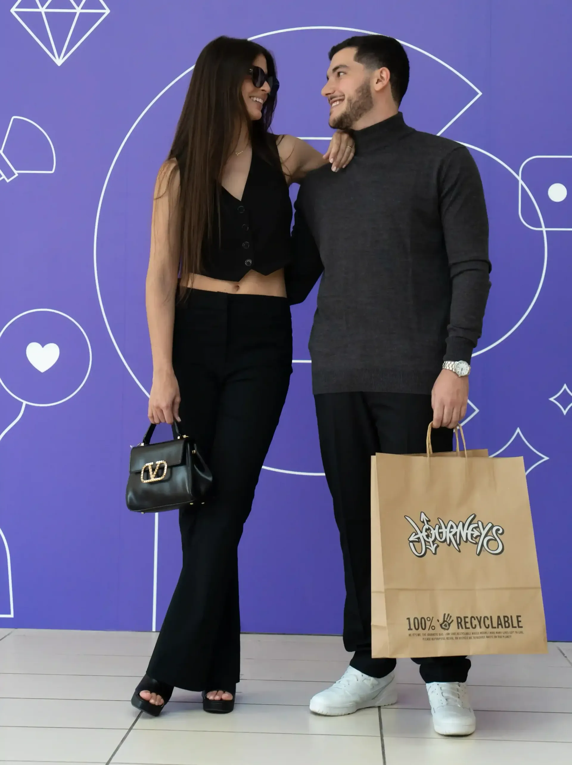 A stylish couple poses in front of a purple wall with white graphics. Céntrico Guayama