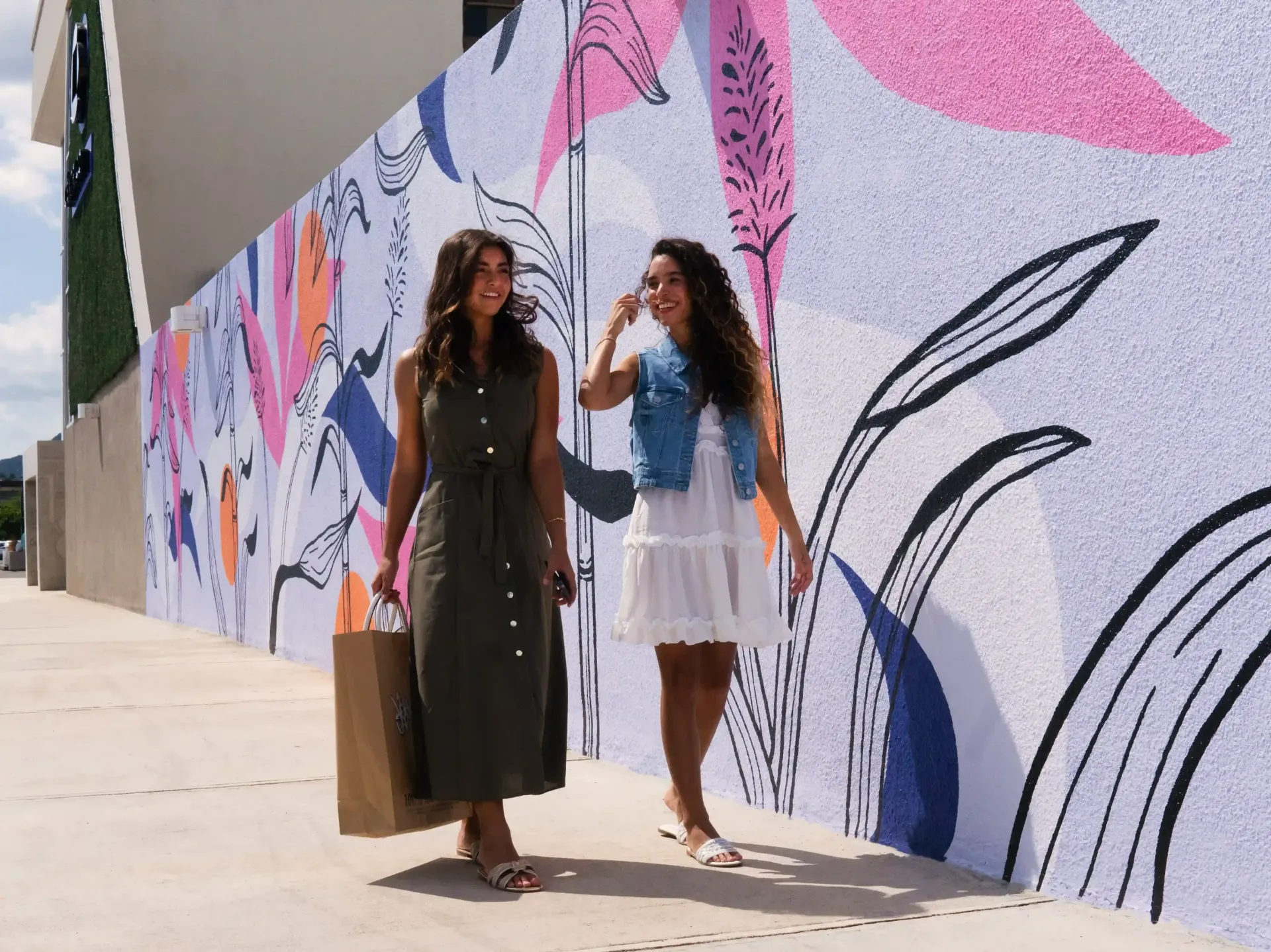 Two women walk past a colorful mural, in Céntrico Guayama, one carrying a shopping bag.