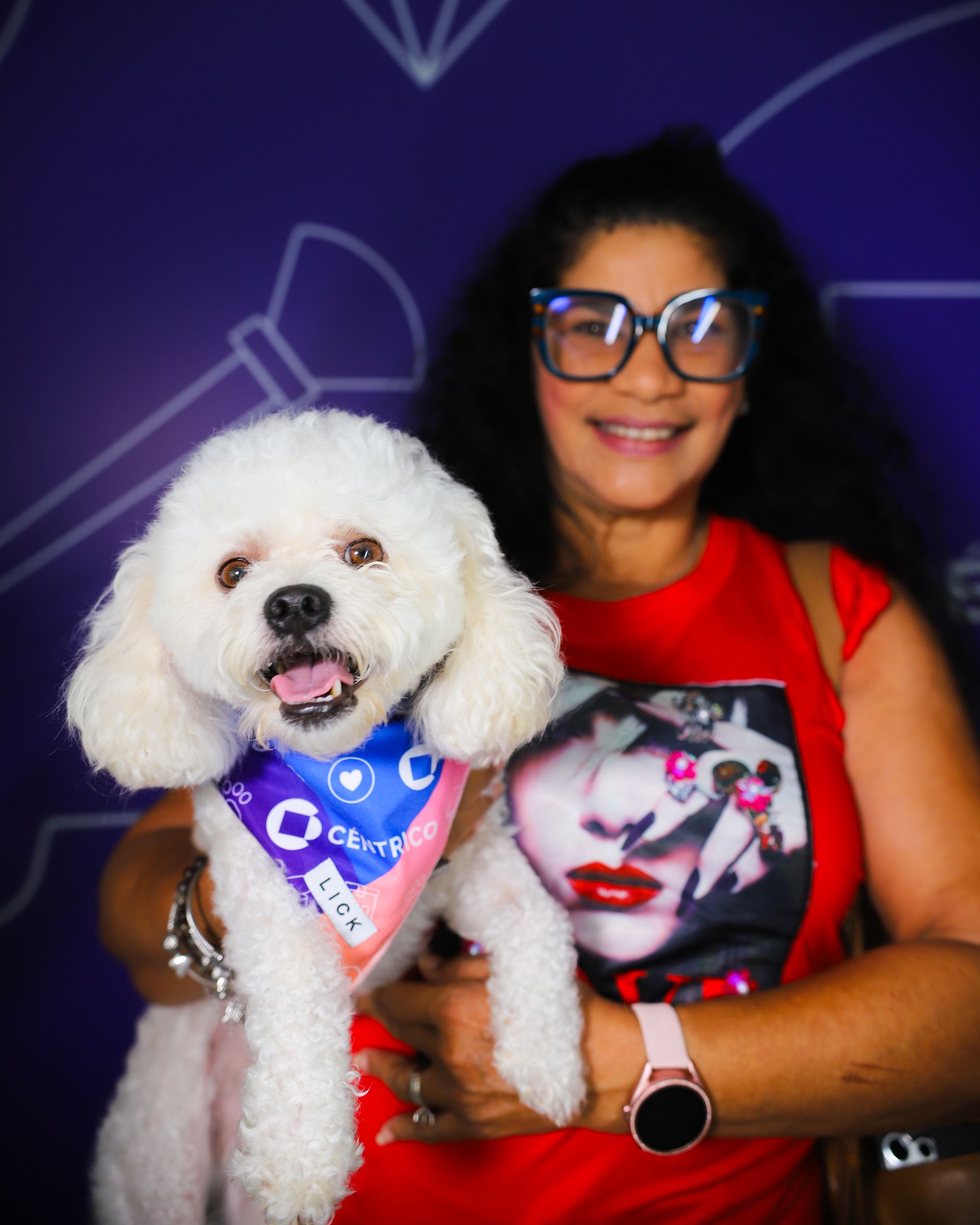Woman holding a fluffy white dog, both smiling. They are in front of a purple background.