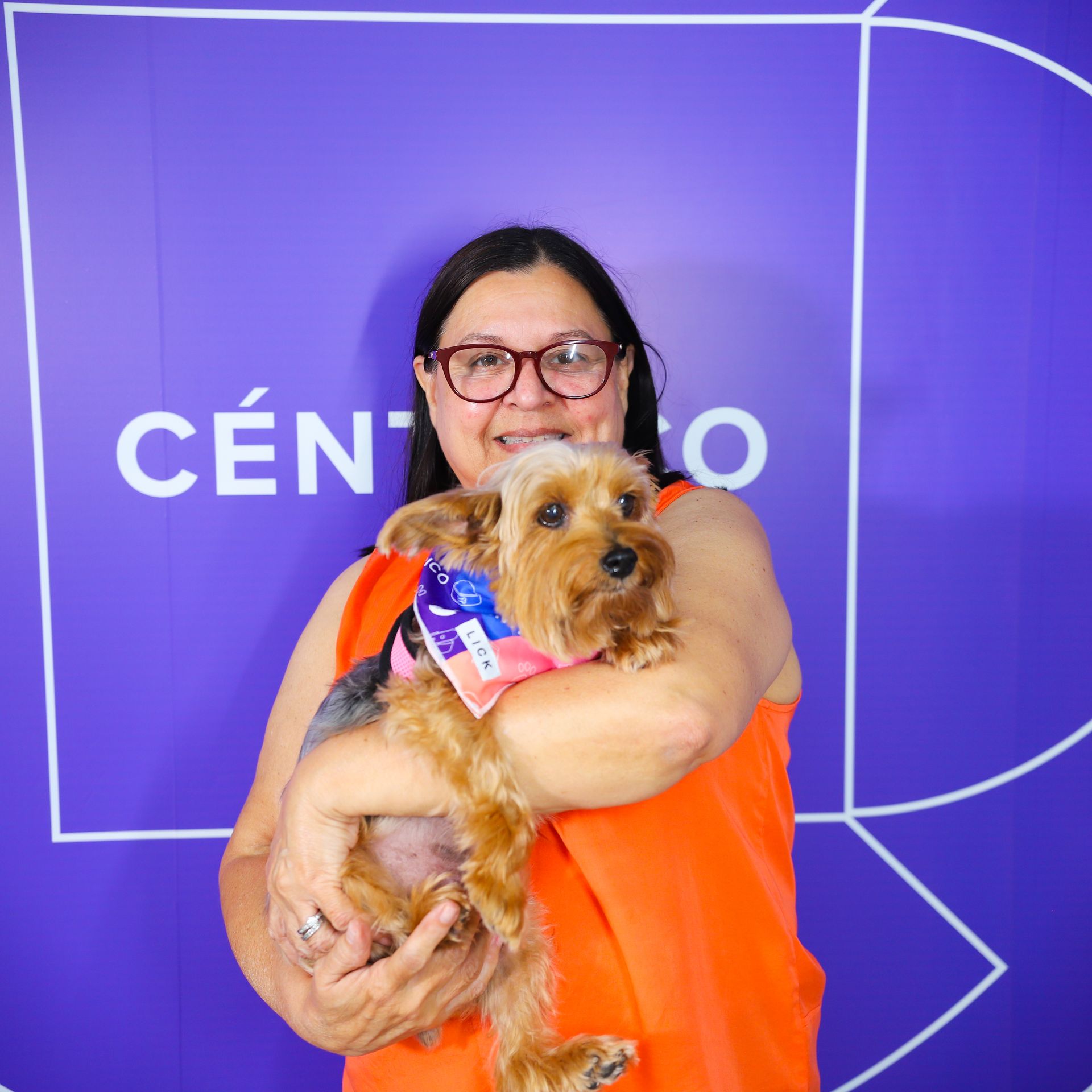 Woman in orange top holds a small dog, both smiling, in front of a purple background.