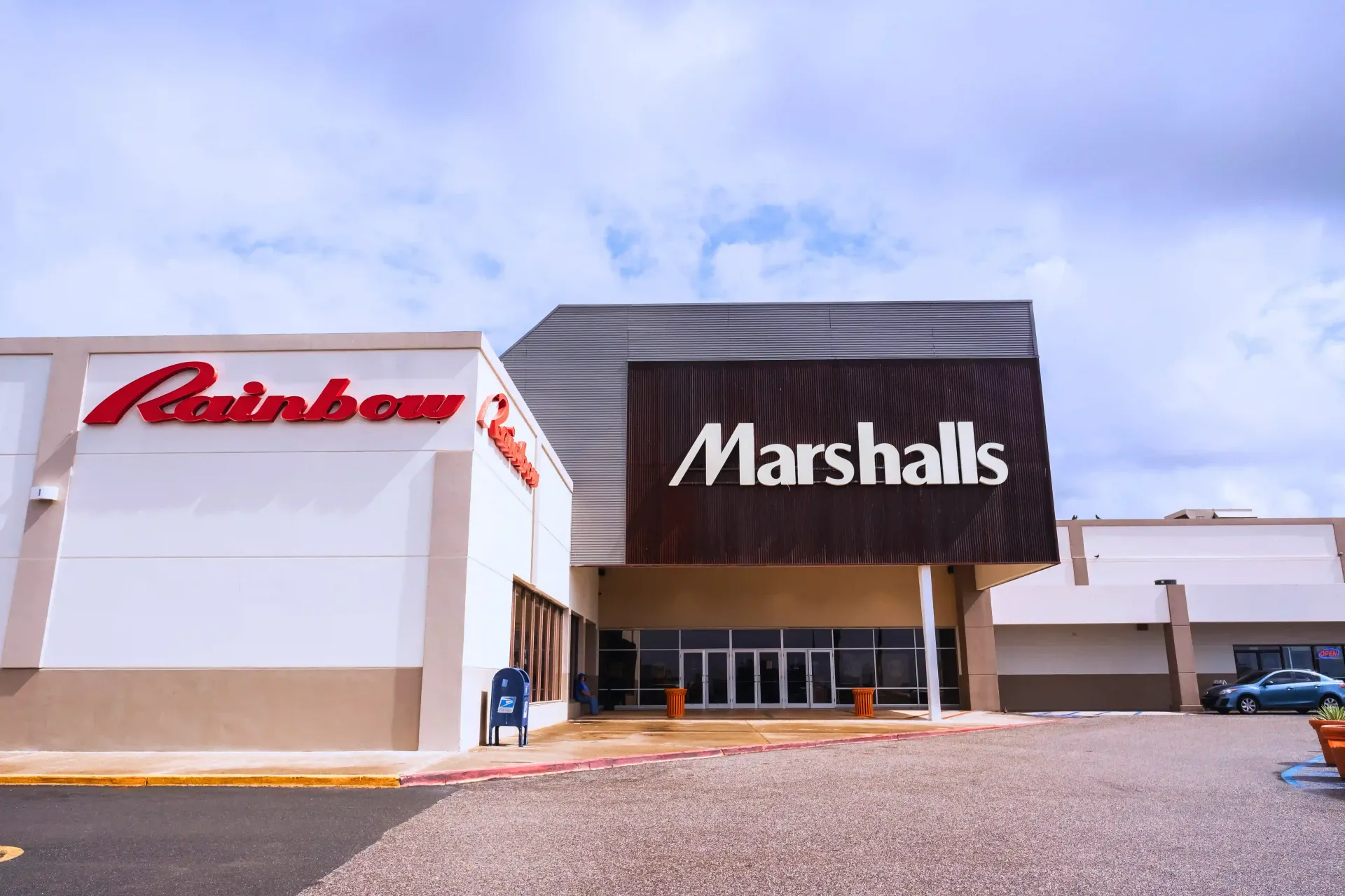 Exterior of a Marshalls store with a Rainbow store on the left, under a cloudy sky.