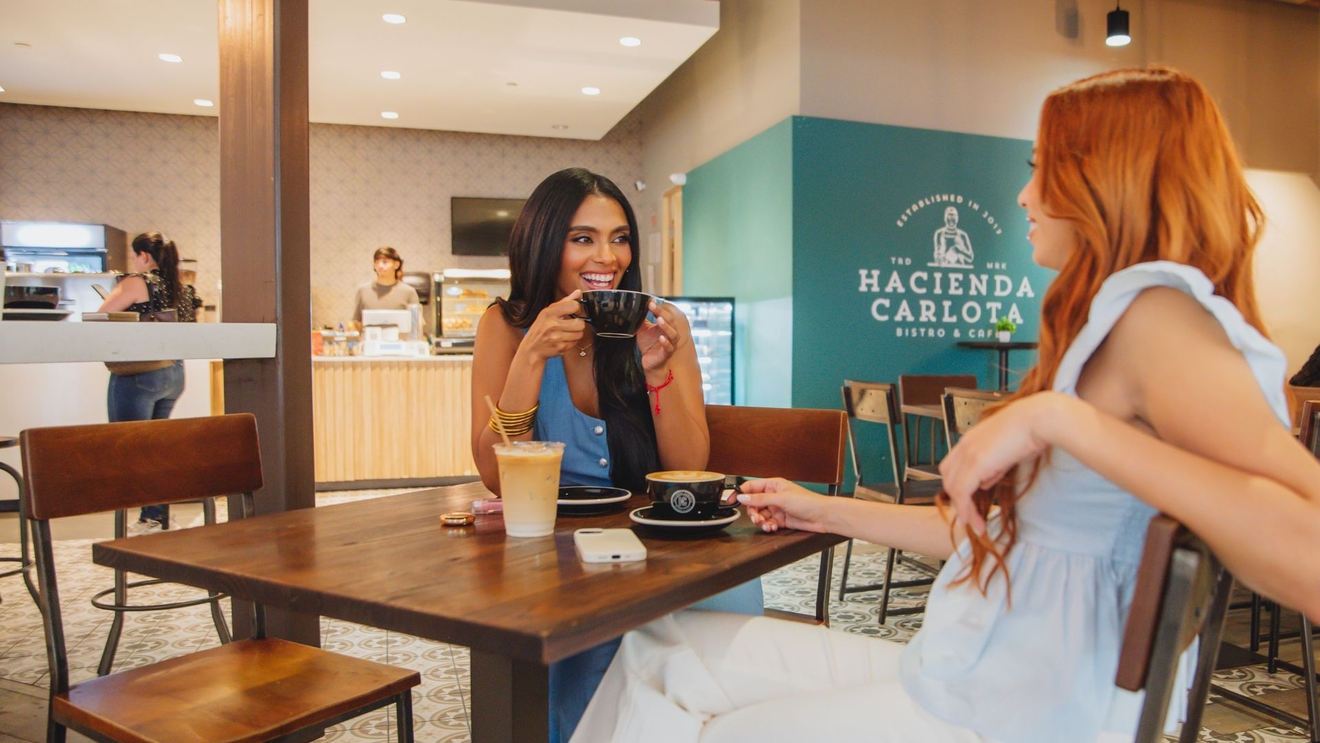 Two women in a cafe, one drinking coffee, smiling. Cafe logo on wall, other people visible.
