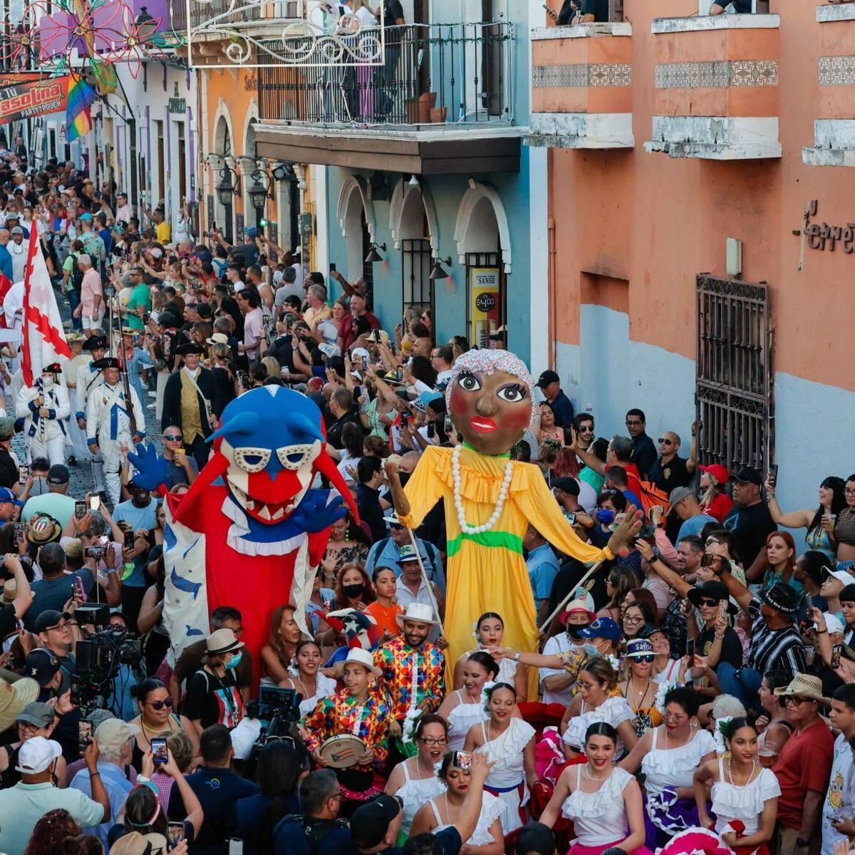 A crowded street festival with costumed figures and a large crowd in a colorful urban setting.
