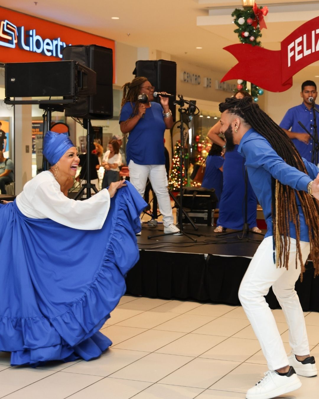 People in blue outfits perform music and dance on a stage in a mall, with holiday decorations.