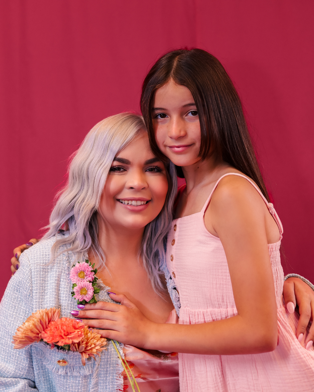 Woman and girl smiling in pink dresses against a pink backdrop, holding flowers
