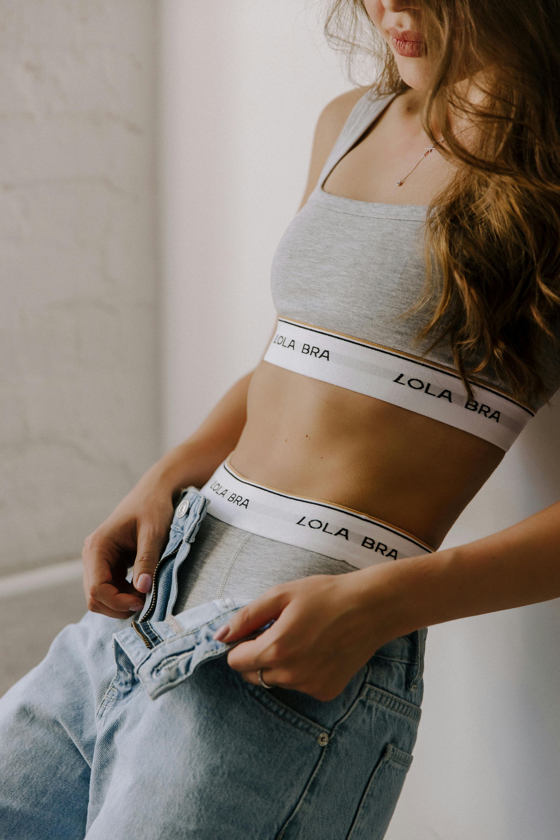 A woman wearing a bra and jeans is standing in front of a wall.