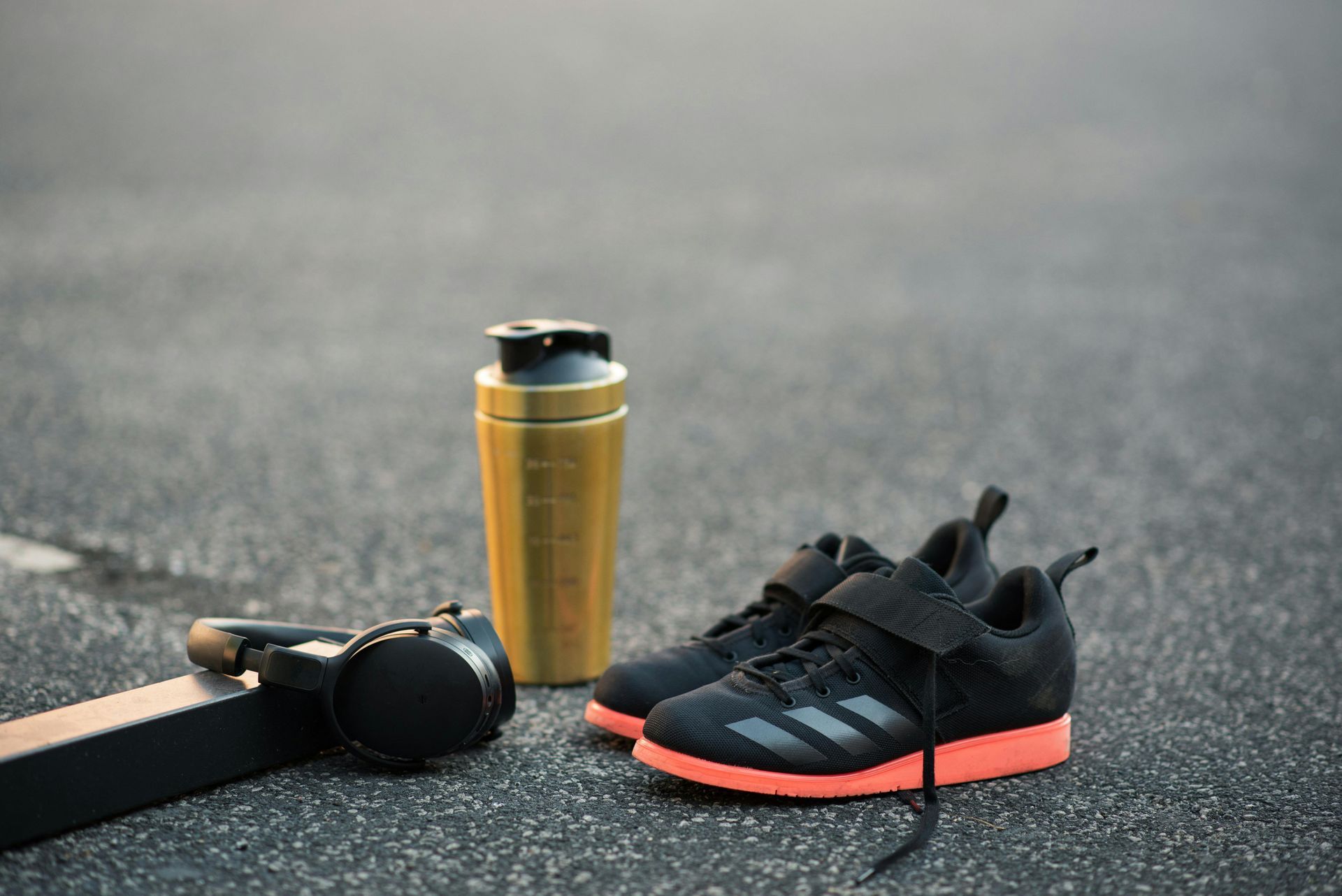 Weightlifting shoes, a gold shaker bottle, and headphones rest on asphalt.