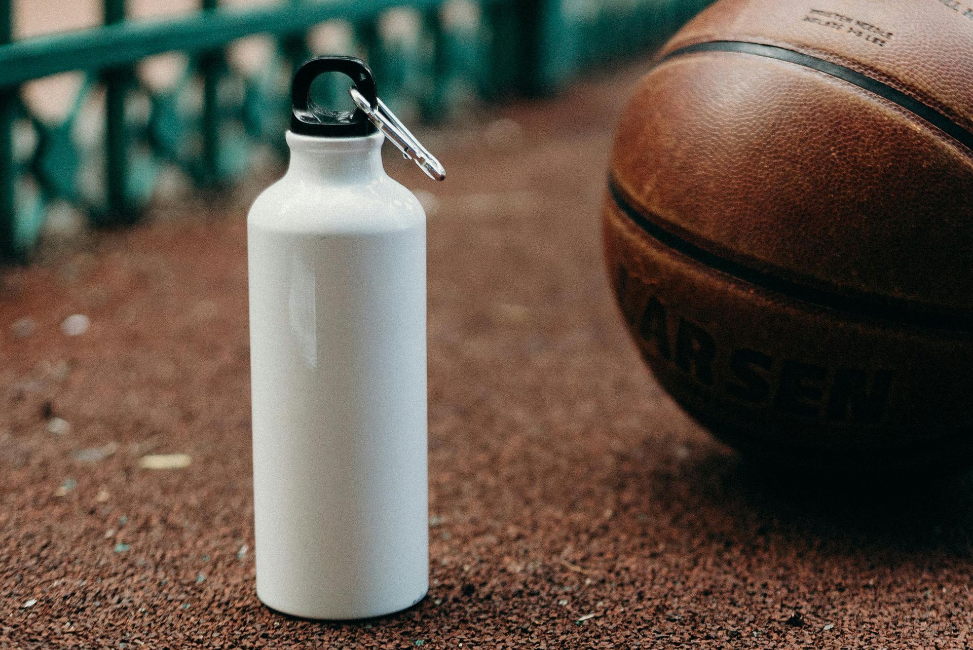 White water bottle next to a brown basketball on a textured brown surface, with a green fence in the blurred background.