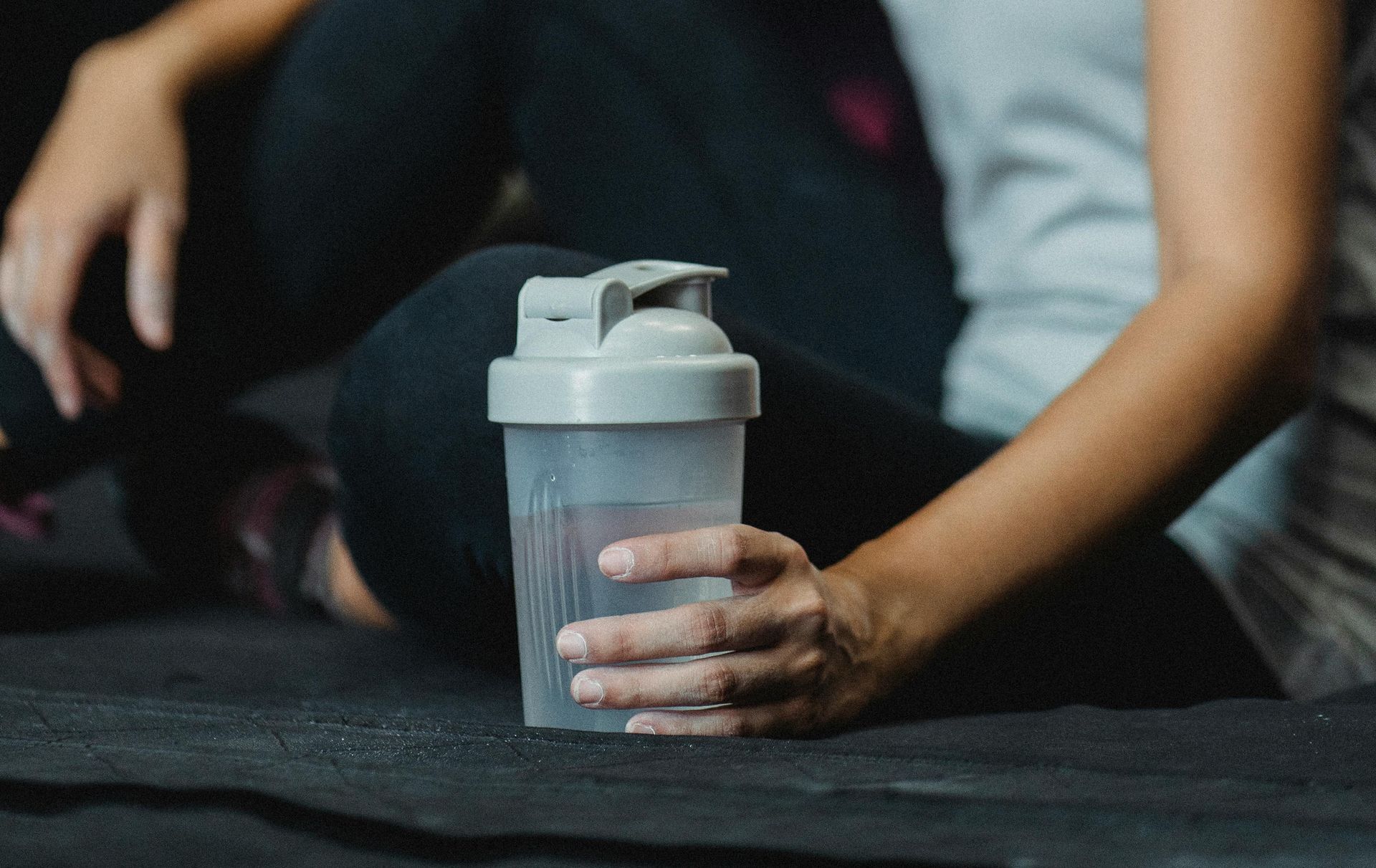 Woman seated, holding a clear plastic water bottle with a grey lid. She is wearing black leggings and a grey tank top.