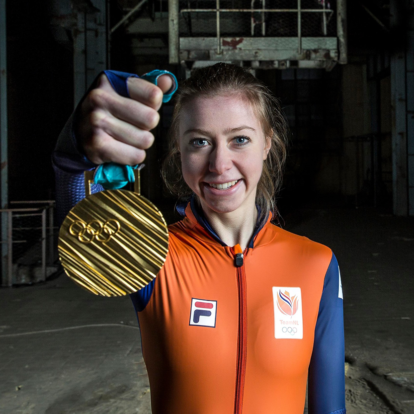 Atleet in oranje jas, met een gouden medaille in de hand, lachend in een industriële omgeving.