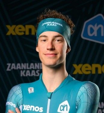 Portret van een lachende atleet in een turquoise shirt en hoofdband met sponsorlogo's op een donkere achtergrond.