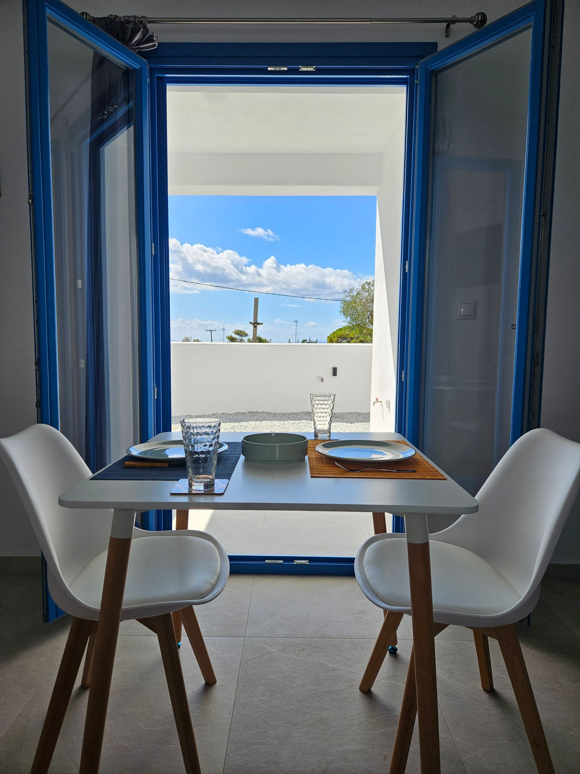 Dining table set for two, framed by blue-trimmed open doors with an outdoor view of a bright blue sky and distant buildings.