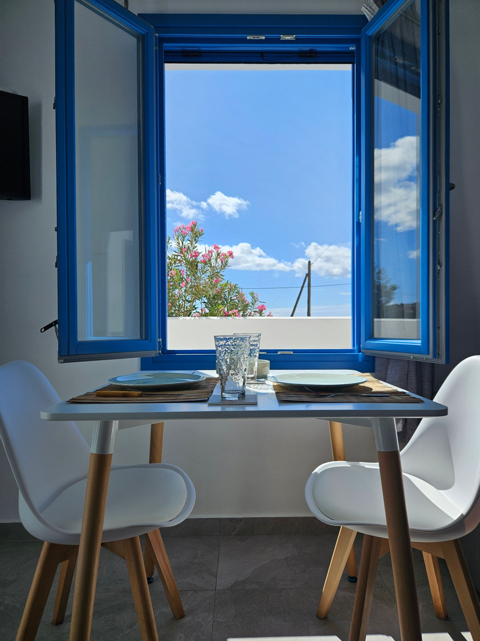 Table set for two in front of a blue-framed window with a view of a bright blue sky and fluffy clouds.
