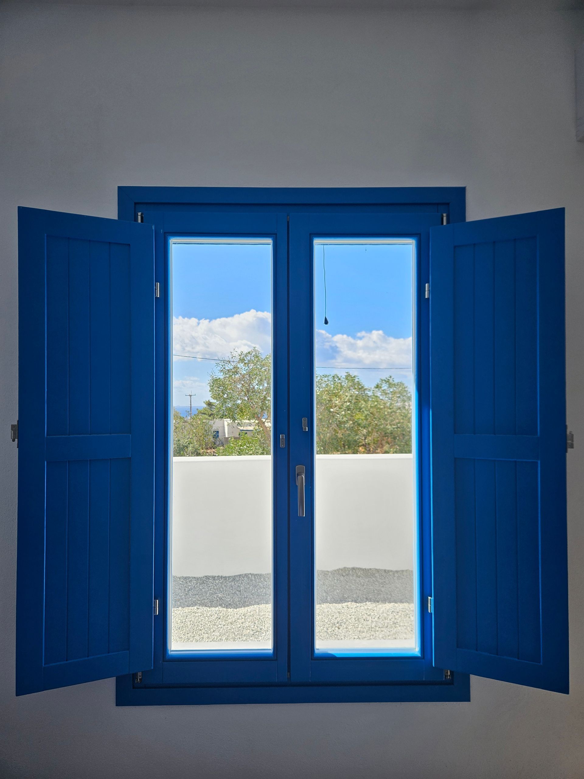 Blue shutters open on a window, revealing blue sky and a white wall, possibly in Greece.